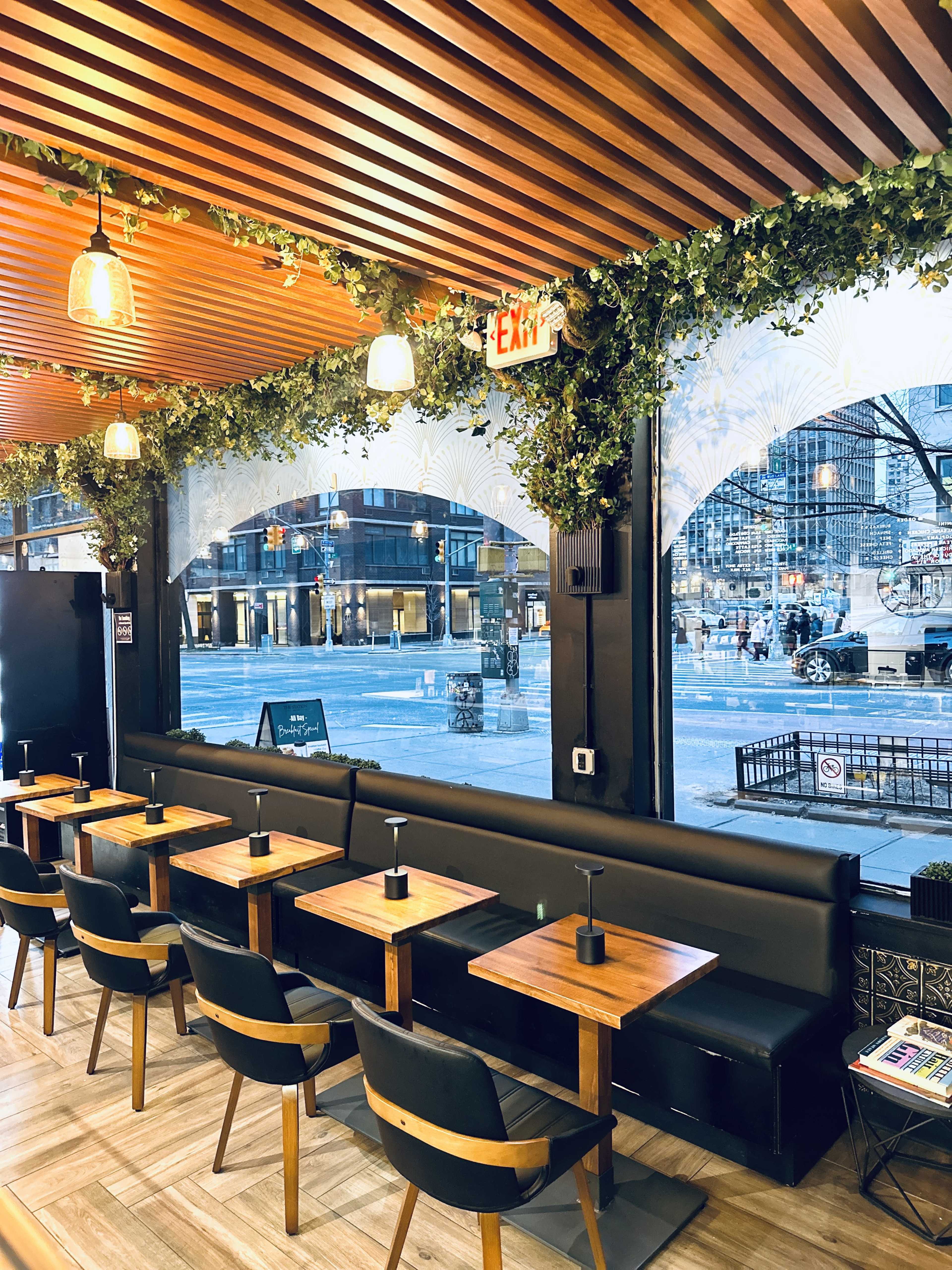 The image shows a modern restaurant interior with wooden tables and black seating, surrounded by large windows and decorative greenery.
