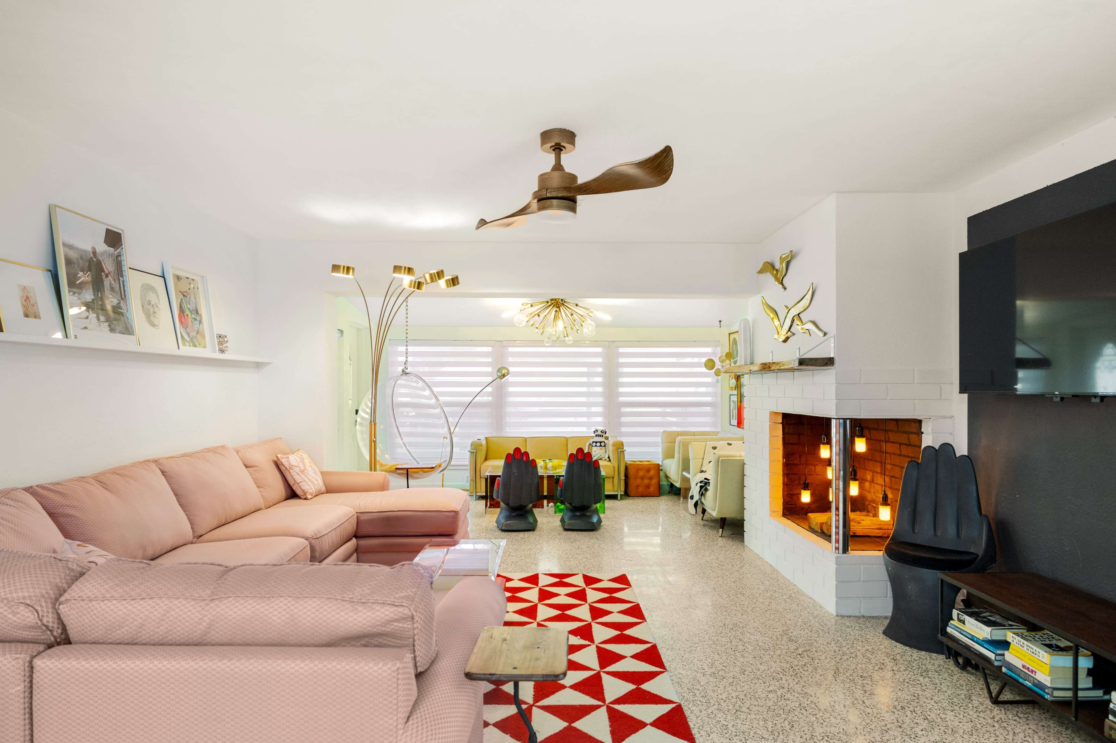 The living room features a pink sofa, a geometric red and white rug, a modern fan, and a fireplace surrounded by white brick, with colorful chairs in the background.