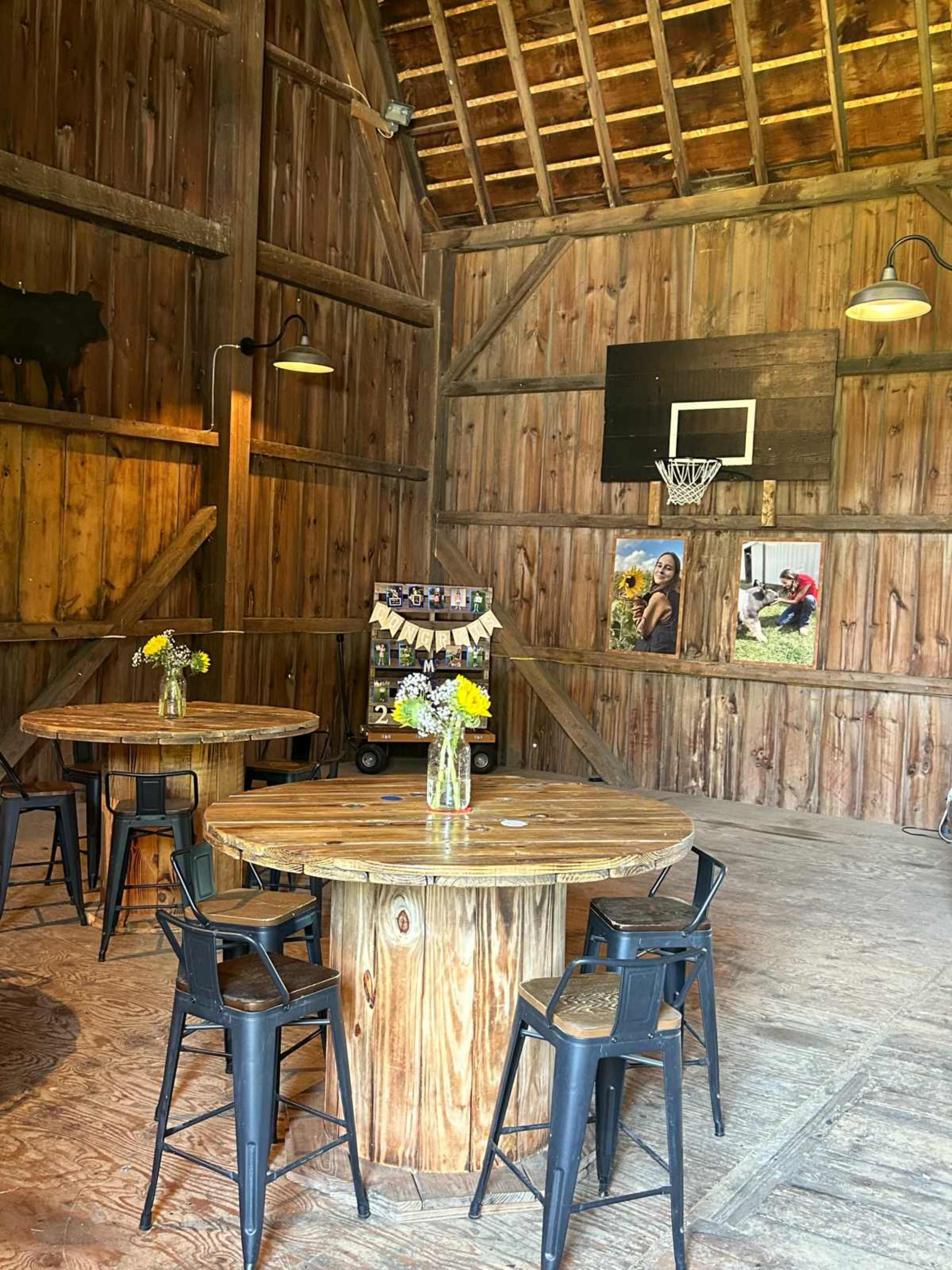 The image shows the interior of a rustic wooden barn featuring two round wooden tables surrounded by black metal chairs, along with a basketball hoop and photographs on the walls.