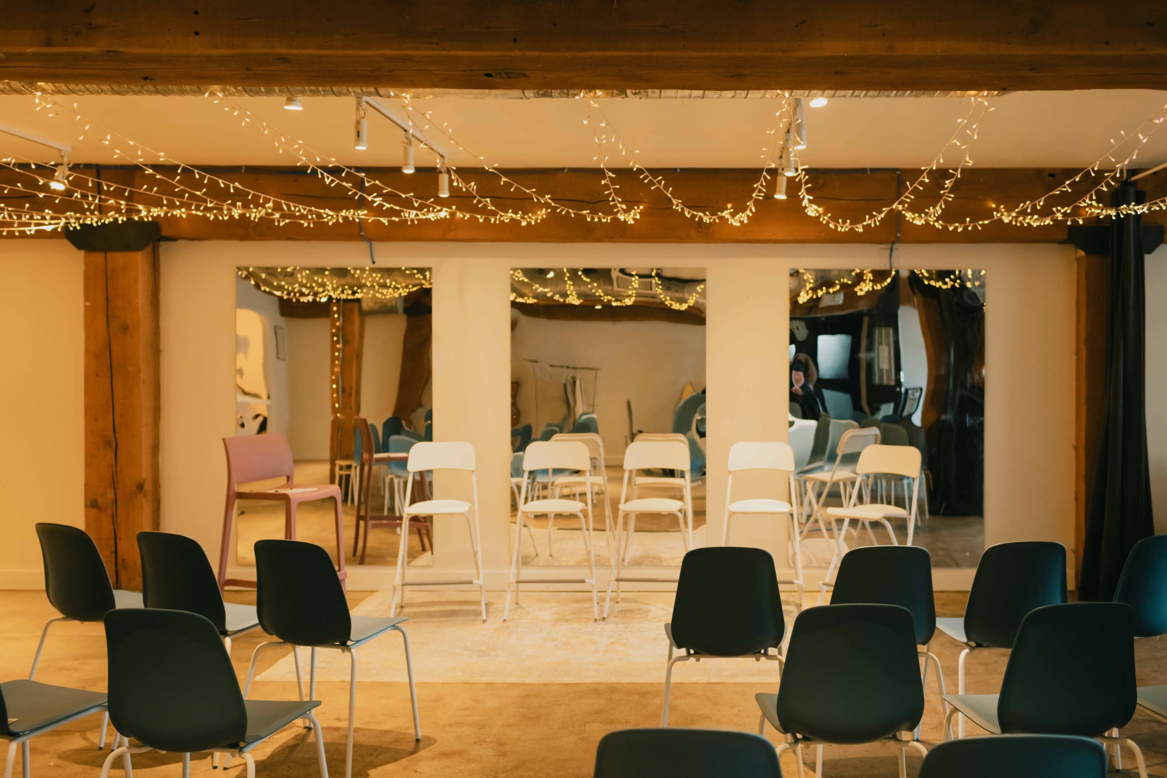 The image shows a carpeted room arranged for a meeting or event, featuring rows of chairs facing a mirrored wall with string lights overhead.
