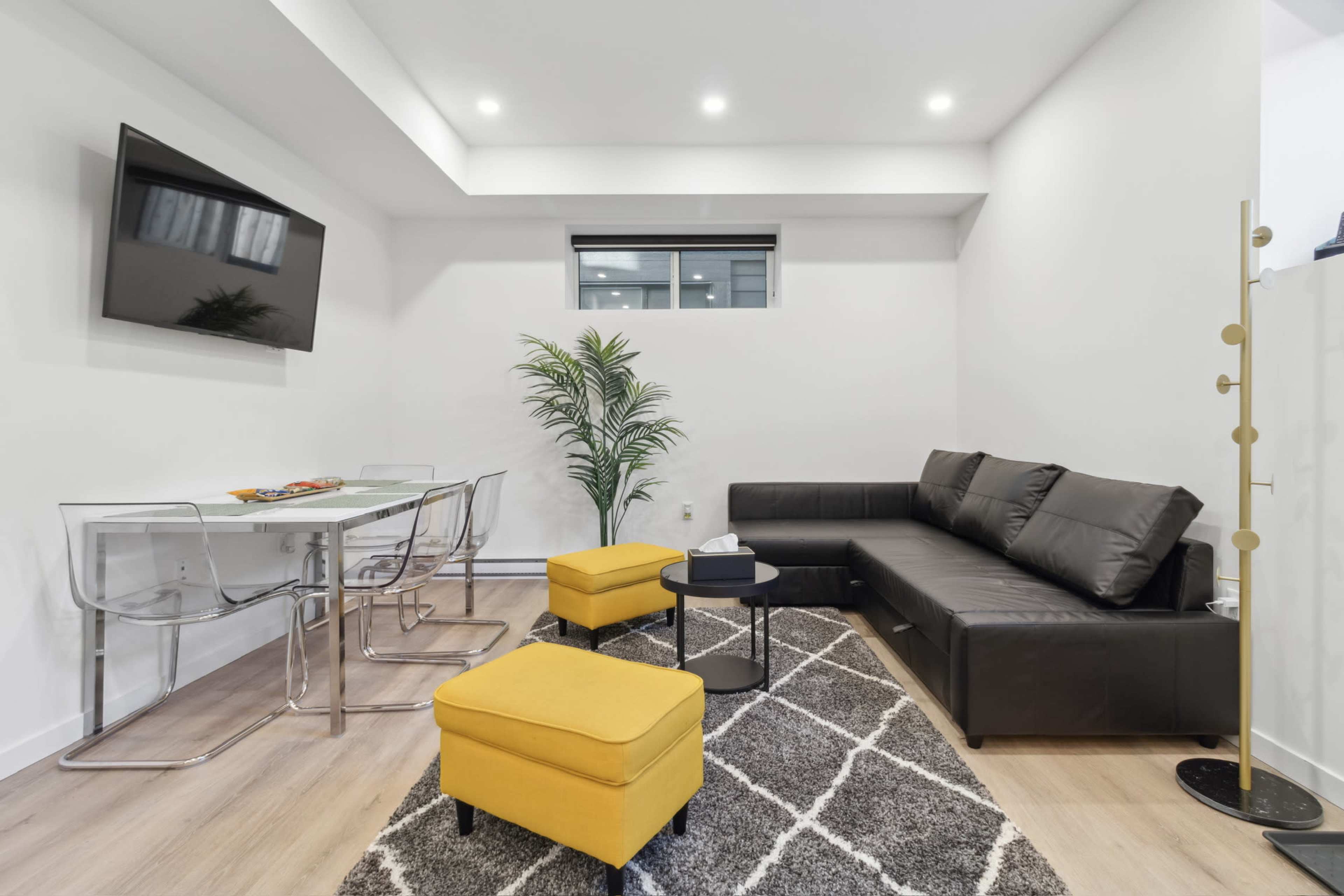 The image shows a modern living space featuring a black sectional sofa, yellow ottomans, a glass dining table, and a tall indoor plant against a white wall.