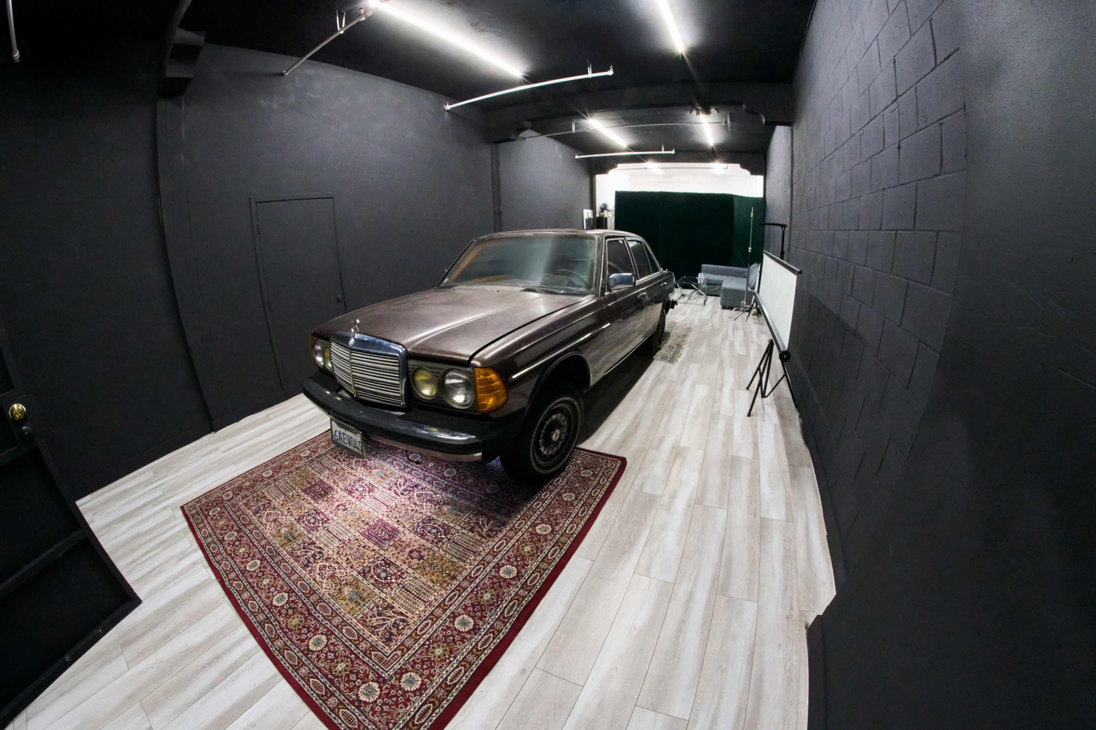 A vintage car is parked on a patterned rug inside a dimly lit room with black walls and wood-like flooring.
