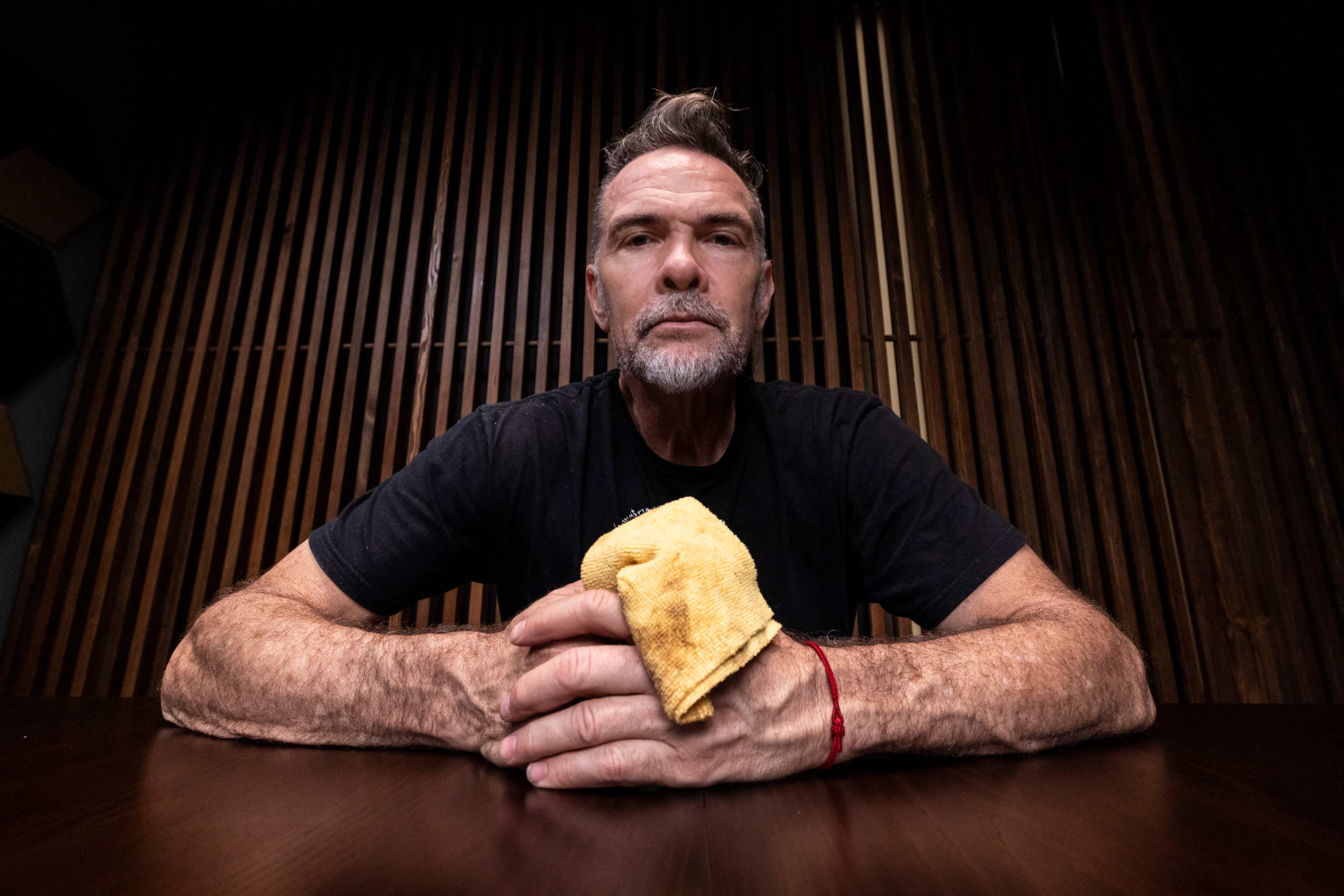 A middle-aged man with a beard sits at a wooden table, holding a yellow cloth in his right hand, against a backdrop of wooden slats.