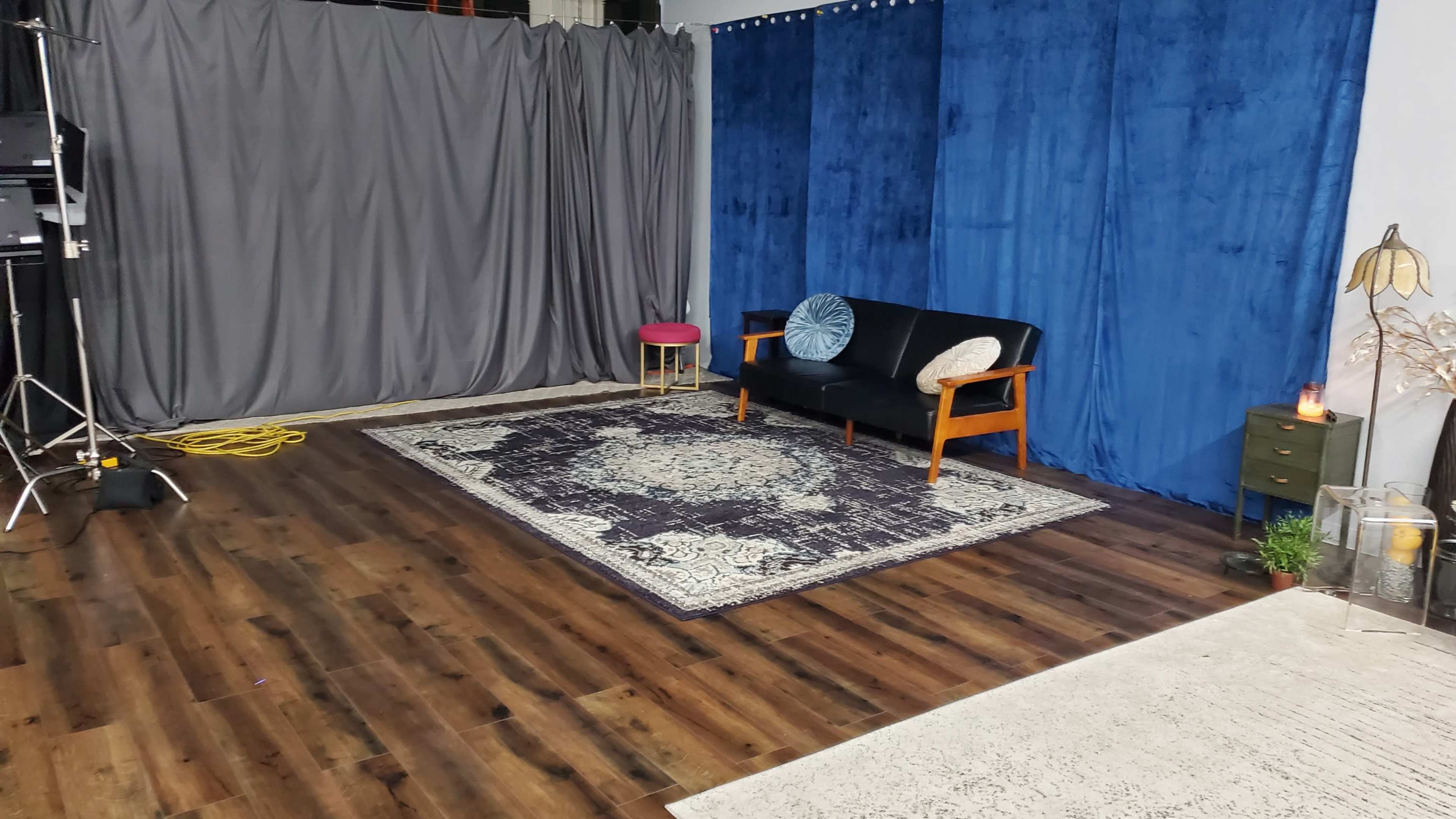 A cozy interior setup with a black sofa, a small pink stool, a patterned rug, and blue and gray fabric backdrops.