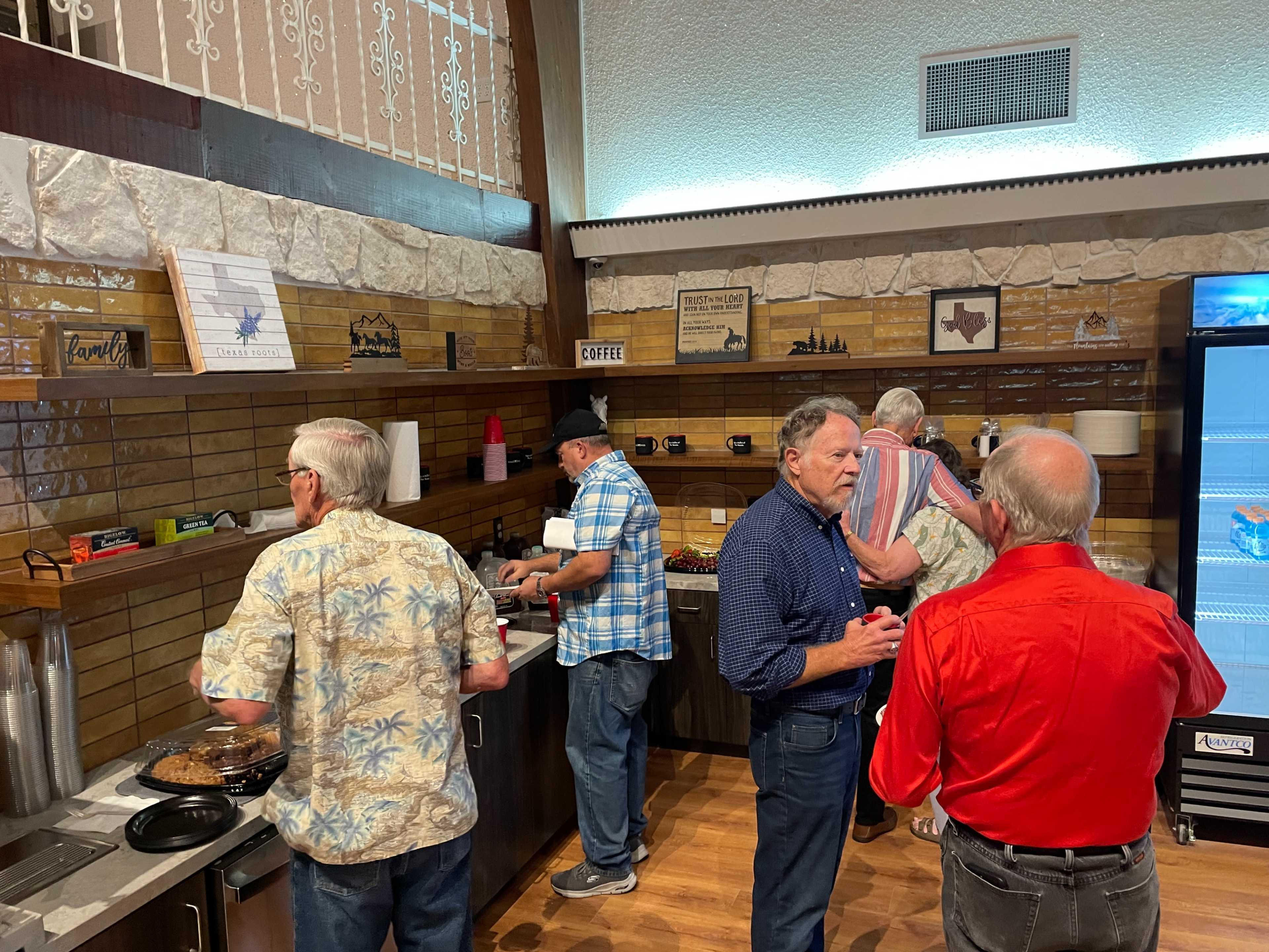 A group of men gather at a snack table in a rustic setting, with one man preparing food while others engage in conversation.