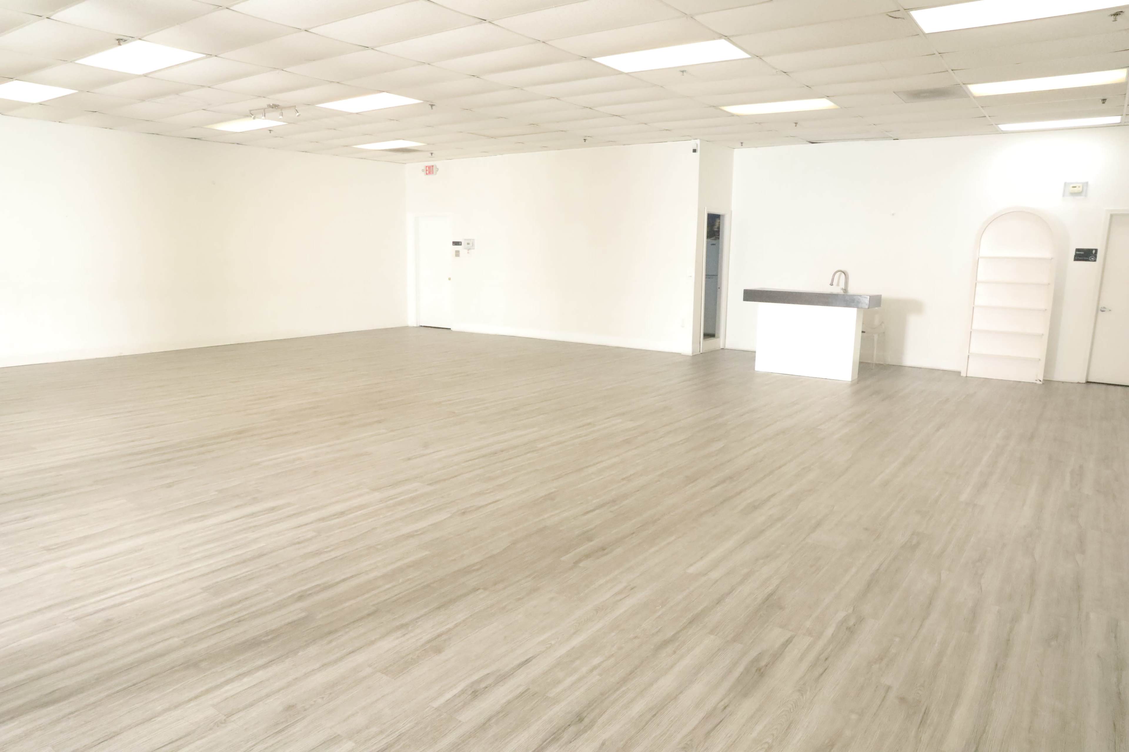 The image shows an empty interior space with light-colored walls and a light wooden floor, featuring a small counter and a built-in shelf at one end.