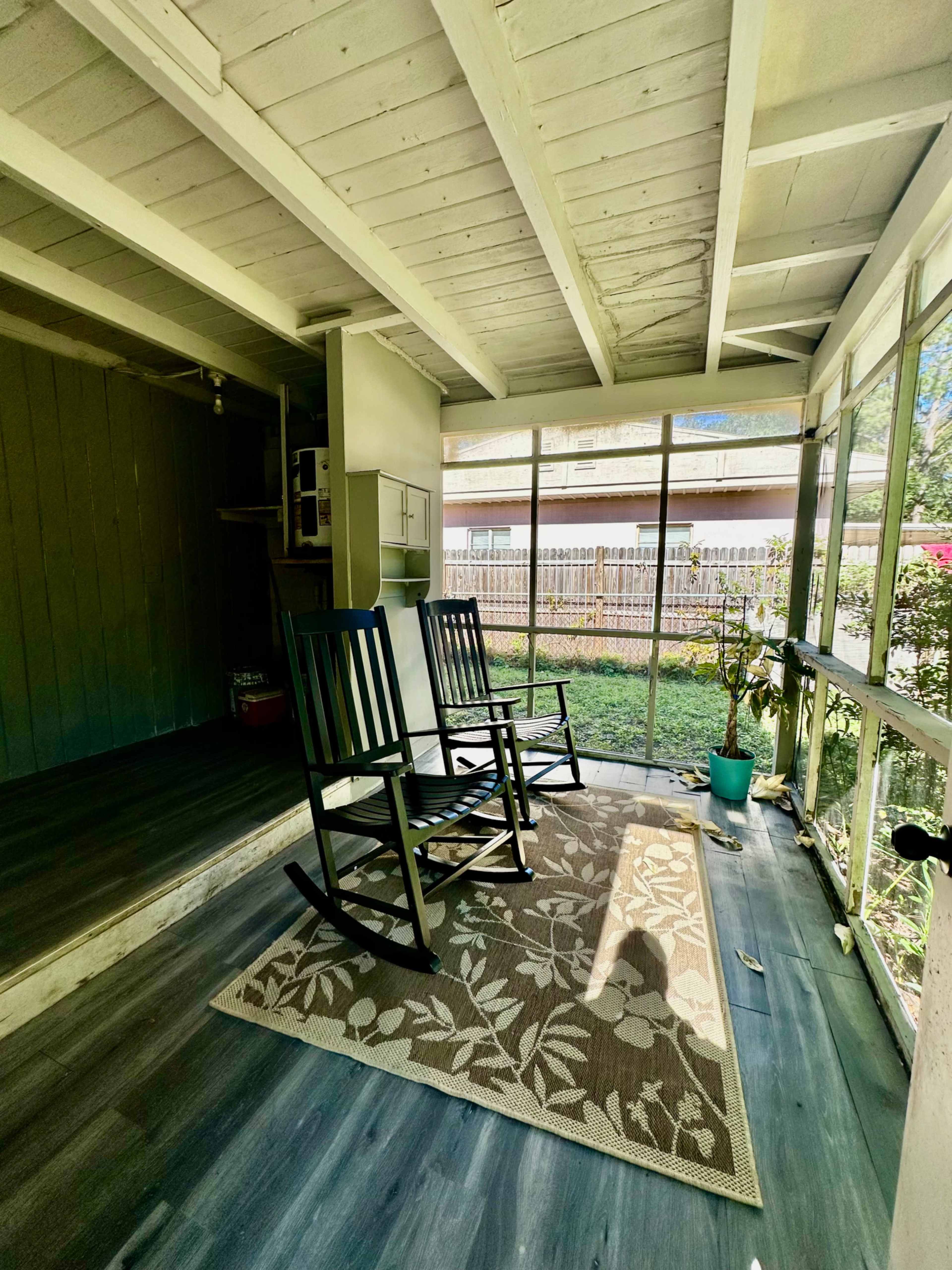 The image shows a screened-in porch with two rocking chairs, a small potted plant, and a decorative rug on the floor.