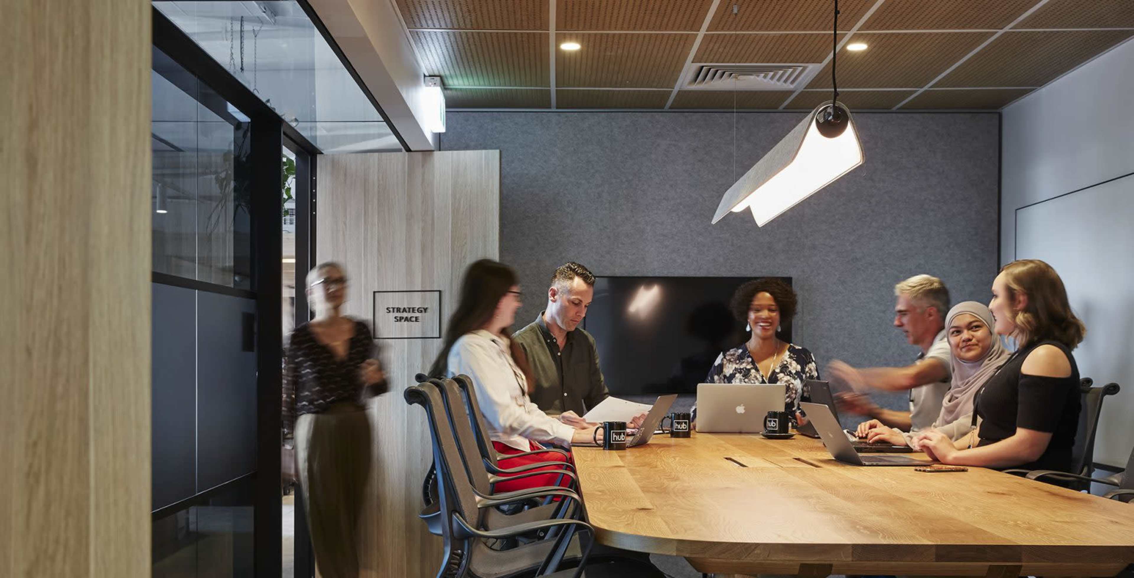 A group of seven people are sitting around a large wooden table in a modern meeting room, engaged in discussion while using laptops and papers.