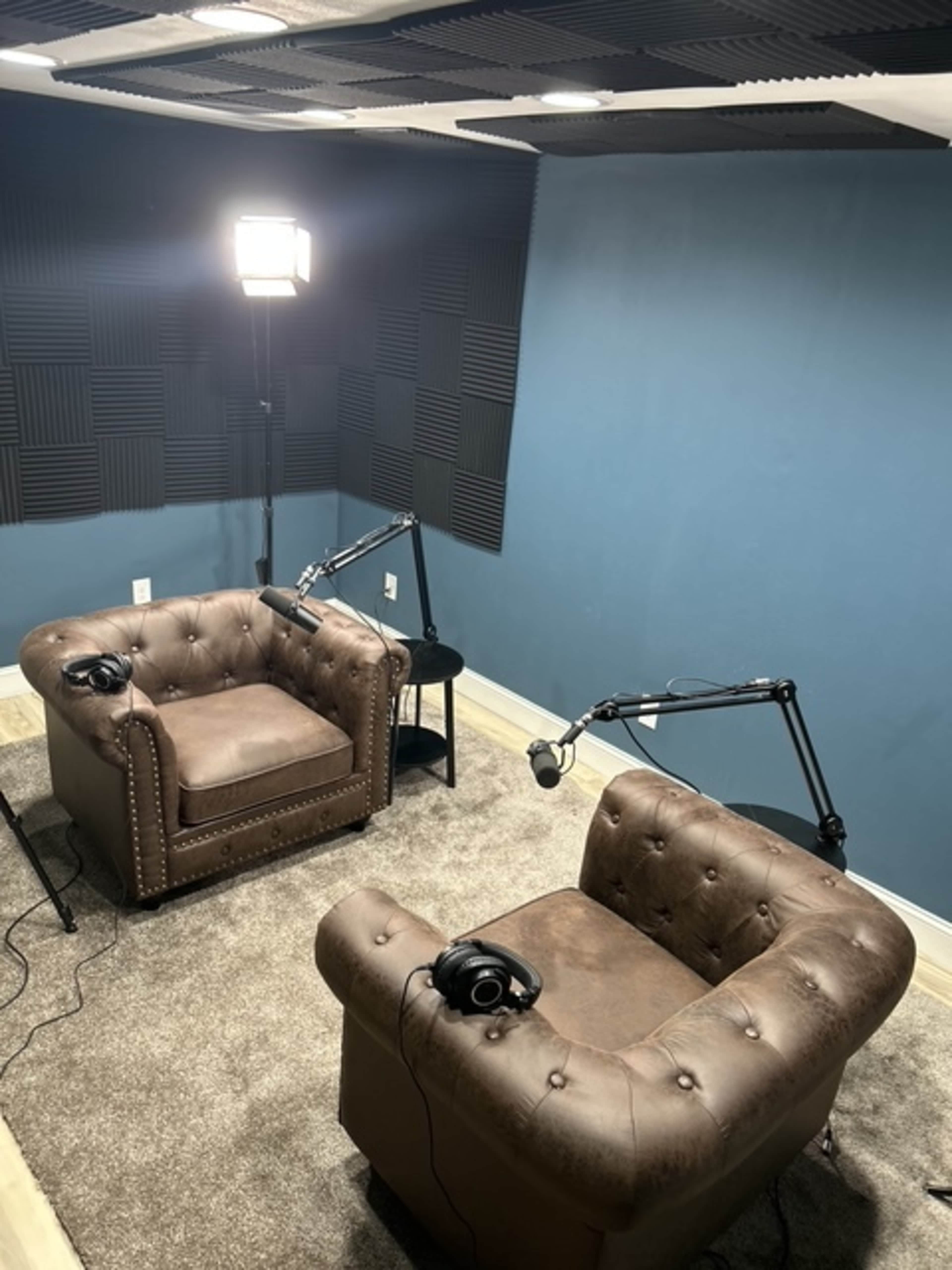 The image shows a small recording studio featuring two brown tufted armchairs, a black side table, and microphone stands, with soundproofing panels on the walls and a light source above.