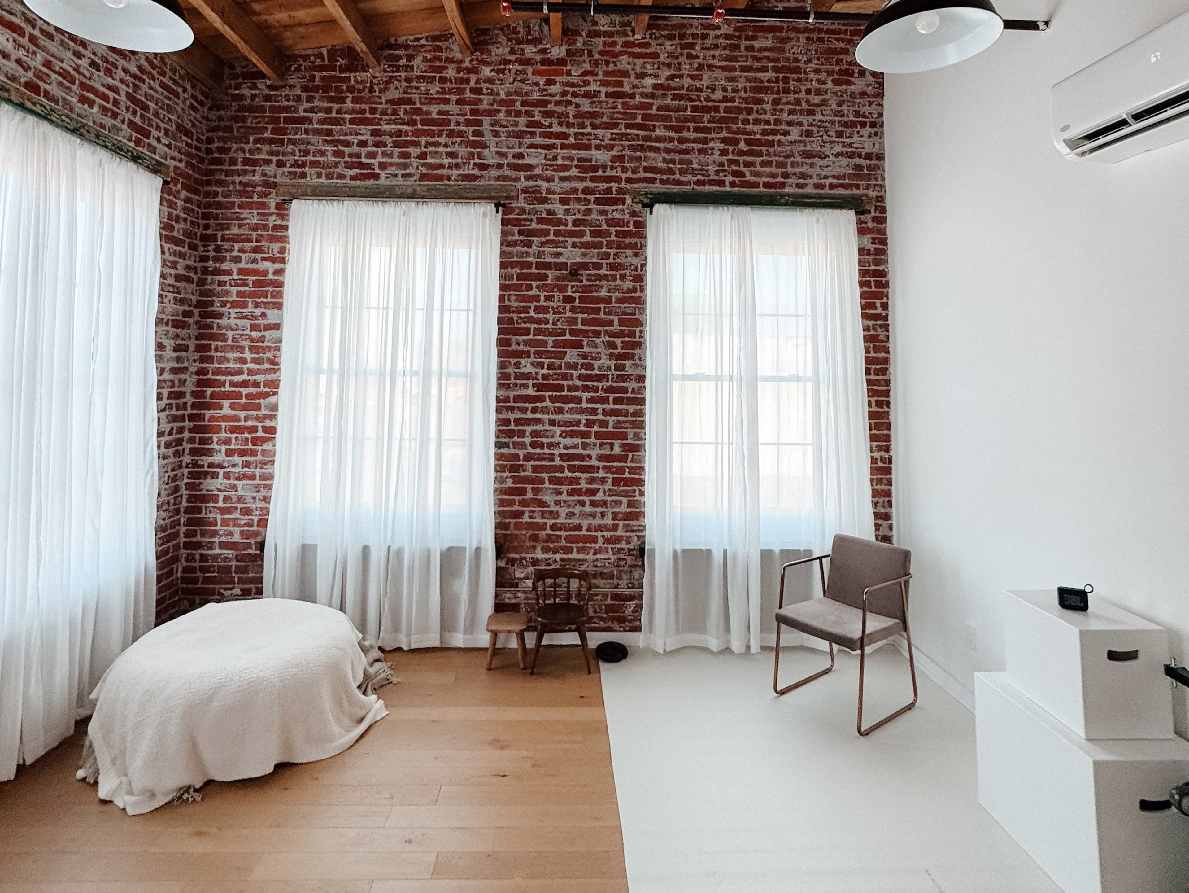 A spacious room features exposed brick walls, large windows with sheer white curtains, a circular bed, a small wooden chair, and a minimalist armchair.