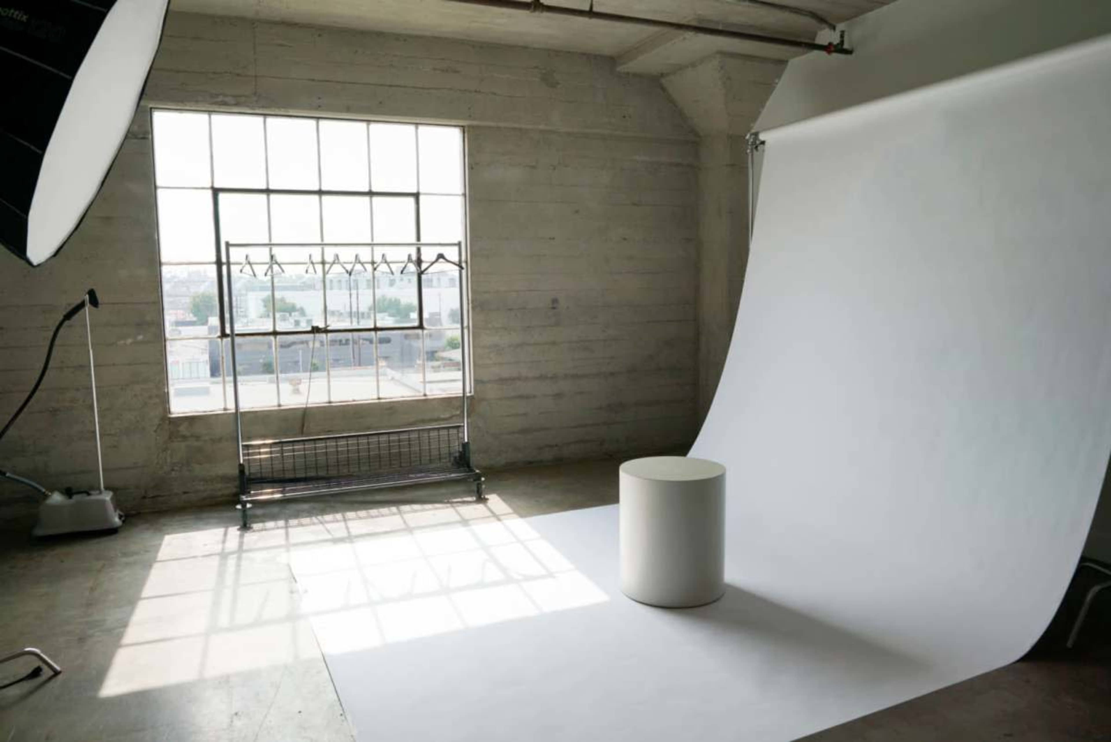 The image shows a photography studio with a large window, a white backdrop, and a round pedestal in the center of the room.