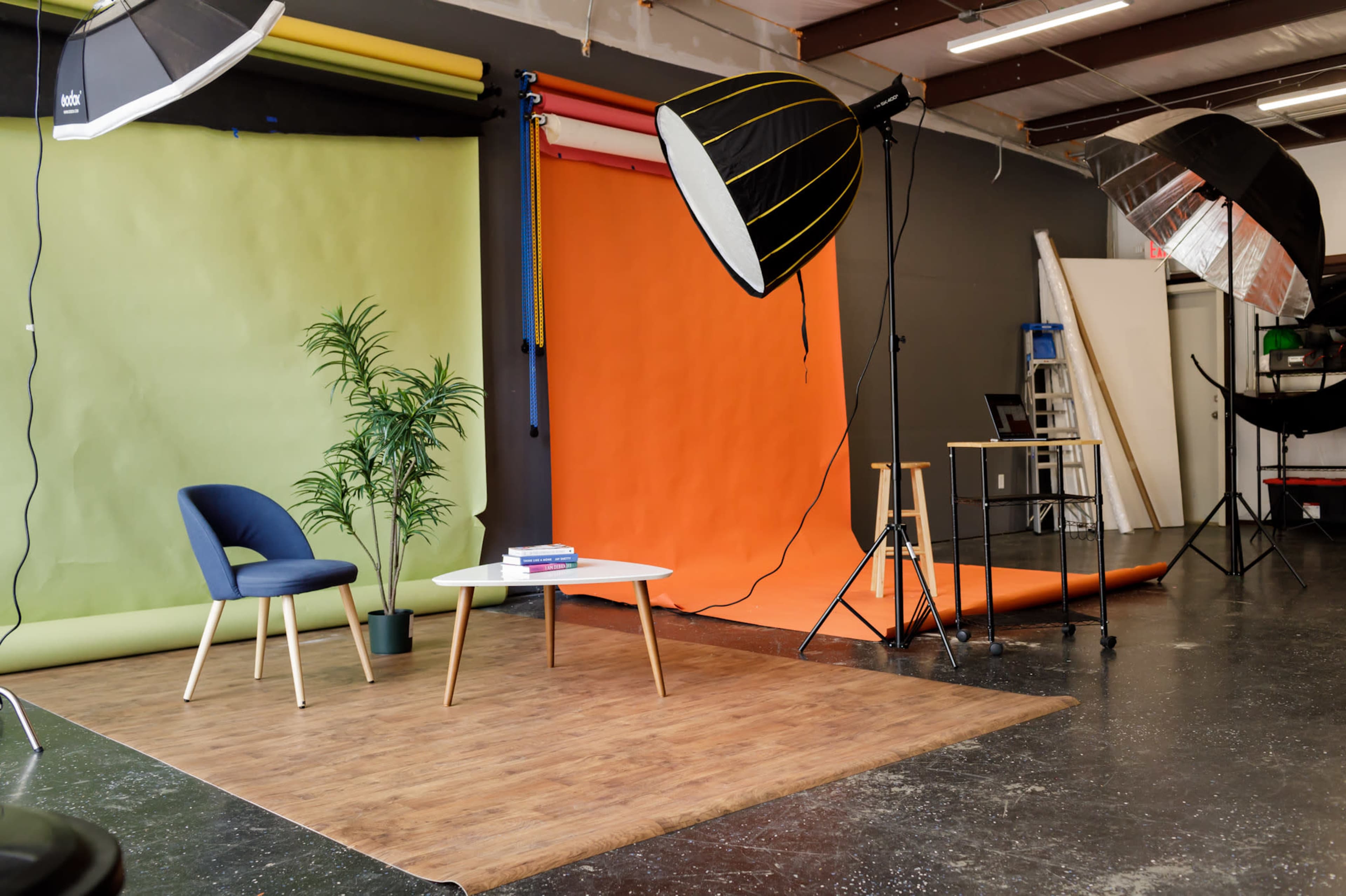 A photography studio with green and orange backdrops, two large lighting setups, a small table with books, a chair, and a plant.