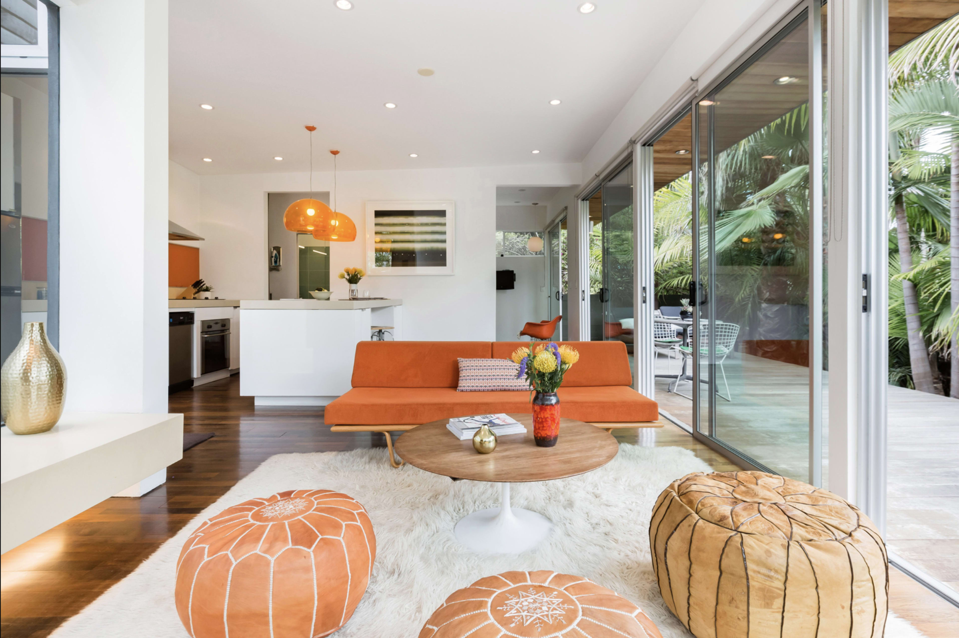 The image shows a modern living space featuring an orange sofa, a wooden coffee table, and decorative poufs, with large glass doors opening to an outdoor area.