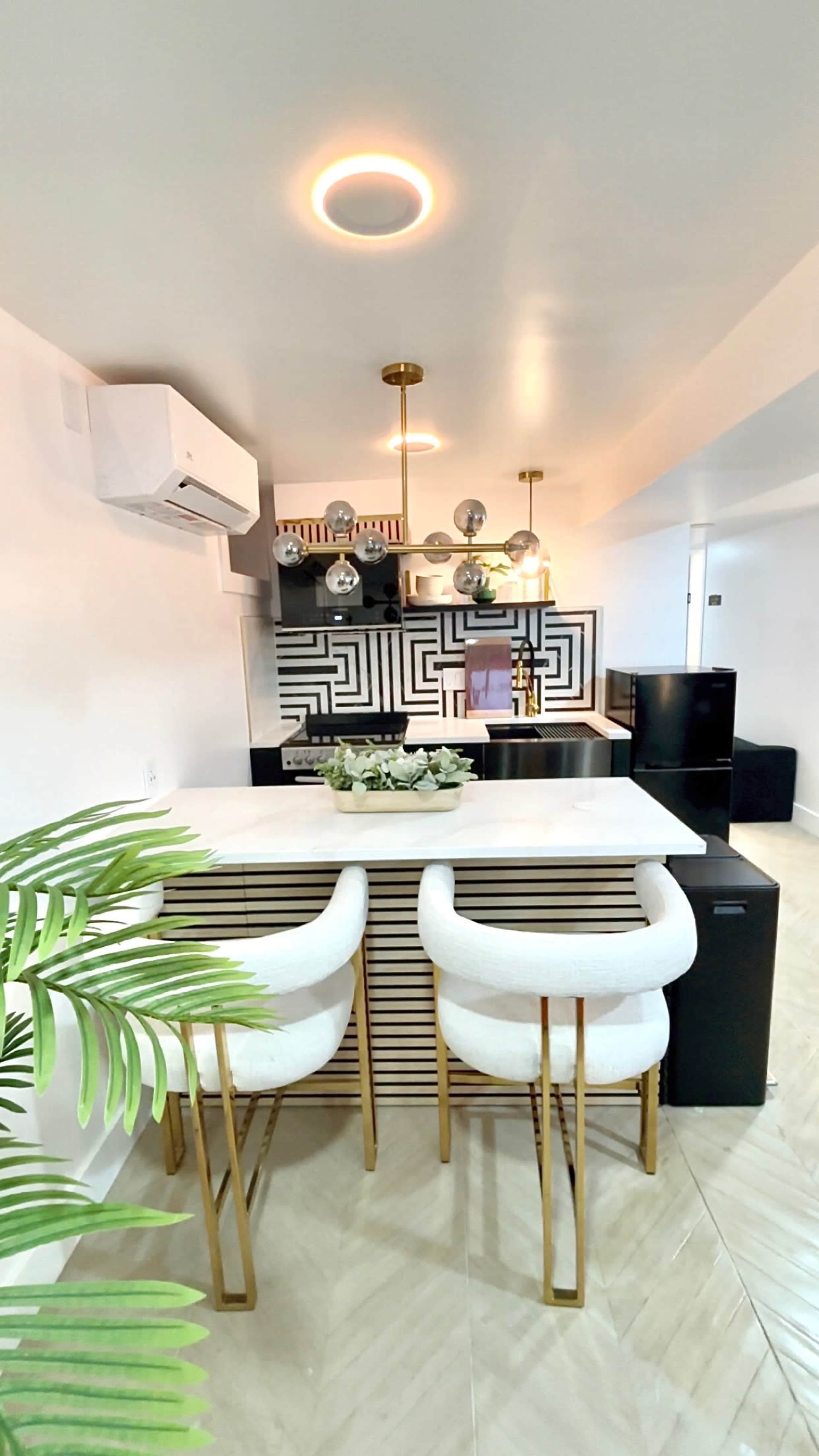 The image shows a modern kitchen with a marble countertop, two bar stools, a decorative backsplash featuring geometric patterns, and overhead lighting fixtures.