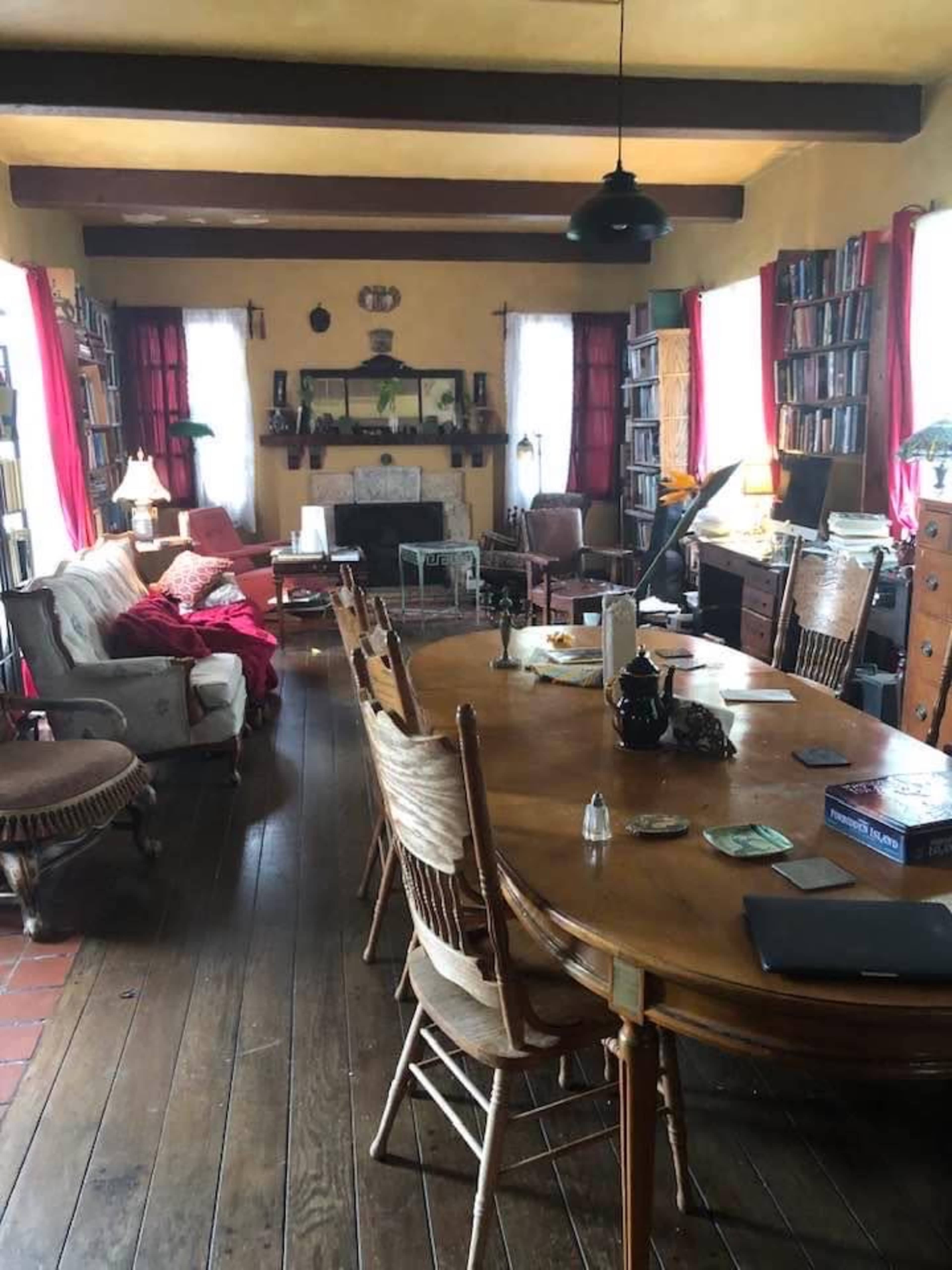 A spacious living room with a large wooden dining table, surrounded by chairs, bookshelves lining the walls, and a mix of furniture in a cozy arrangement.