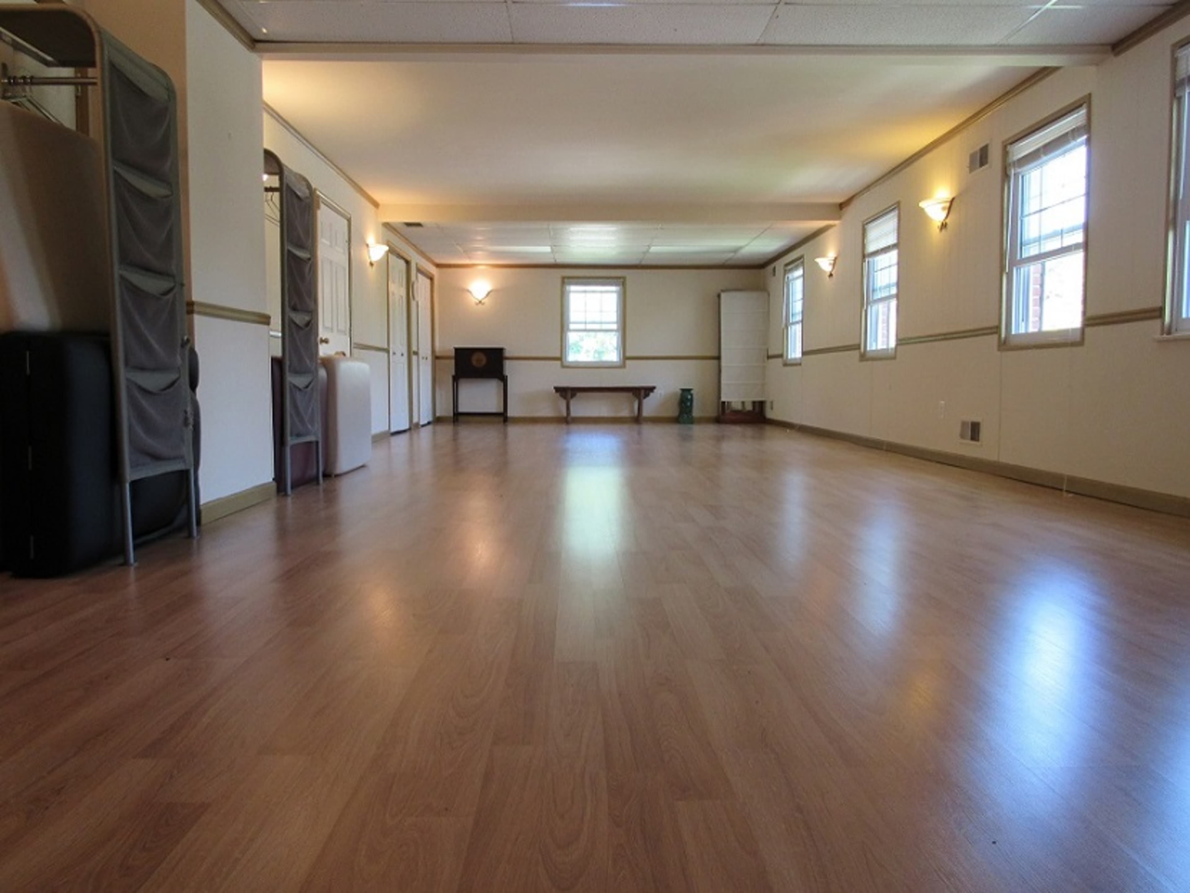 An empty room with wooden flooring, windows on both sides, and a few pieces of furniture along the walls.