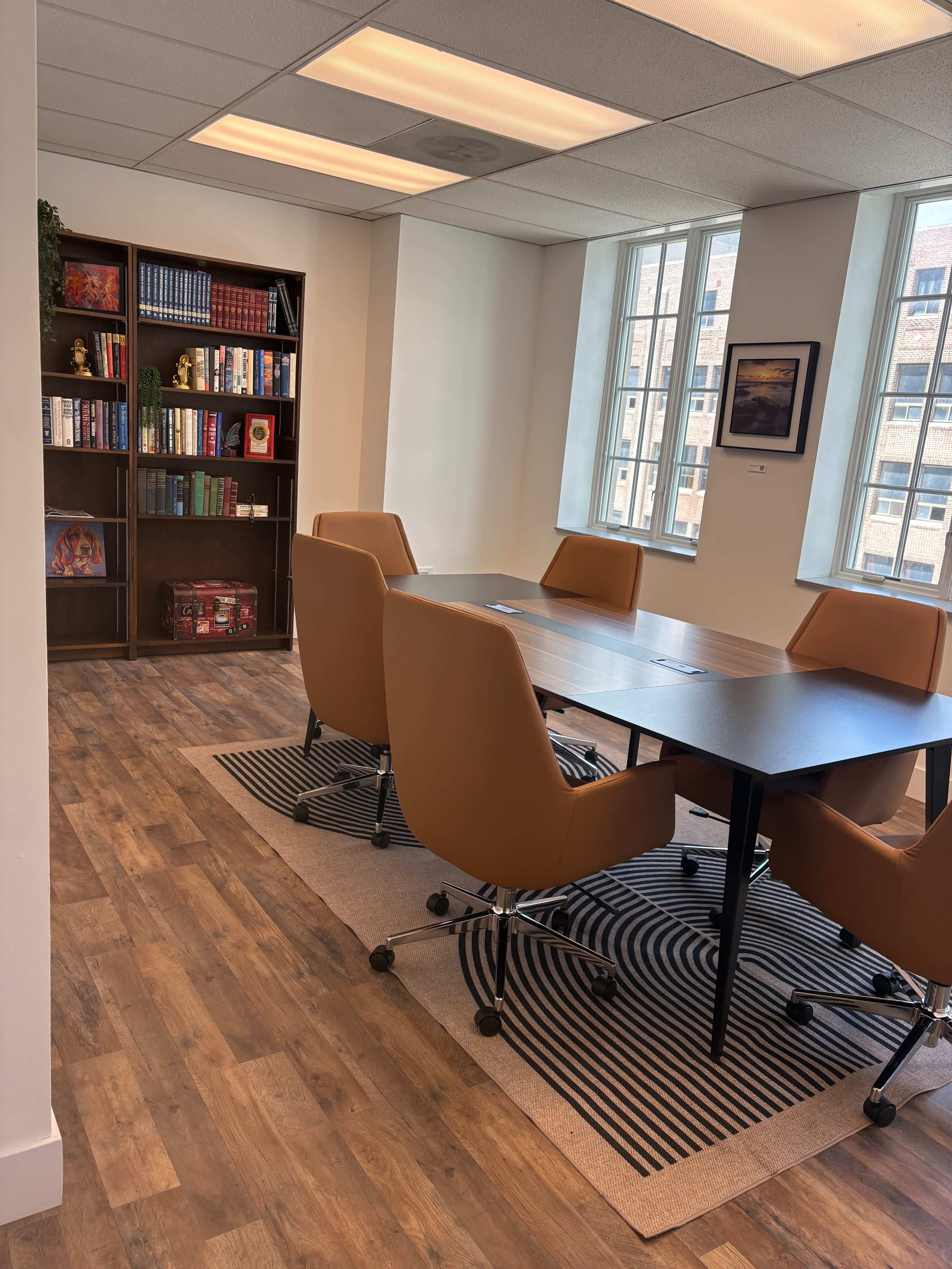 The image shows a bright conference room with a large table surrounded by brown chairs, a bookshelf filled with books, and large windows letting in natural light.