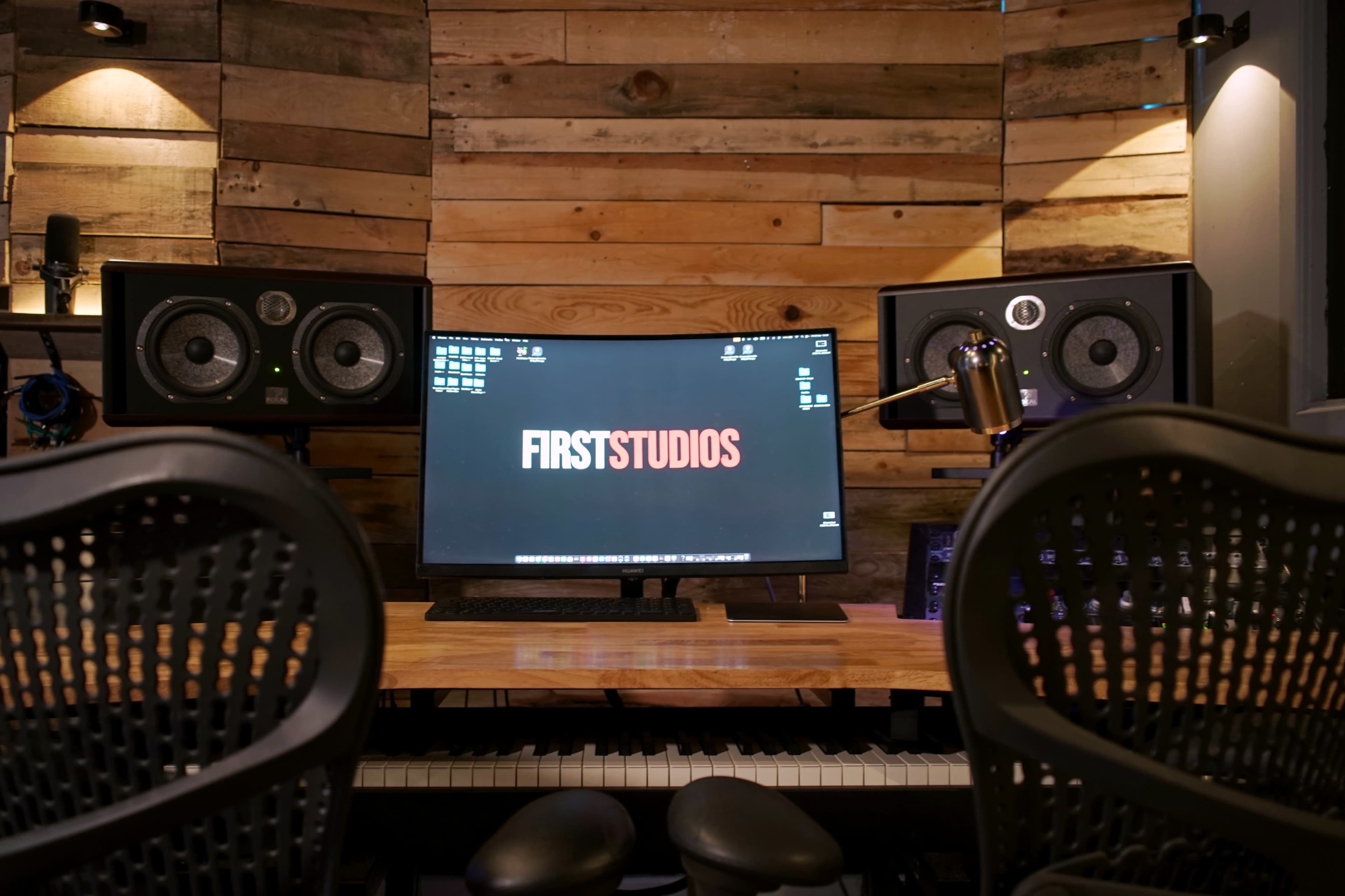 A music production studio features a wooden wall backdrop, a monitor displaying "FIRSTSTUDIOS," and studio speakers flanking the sides.