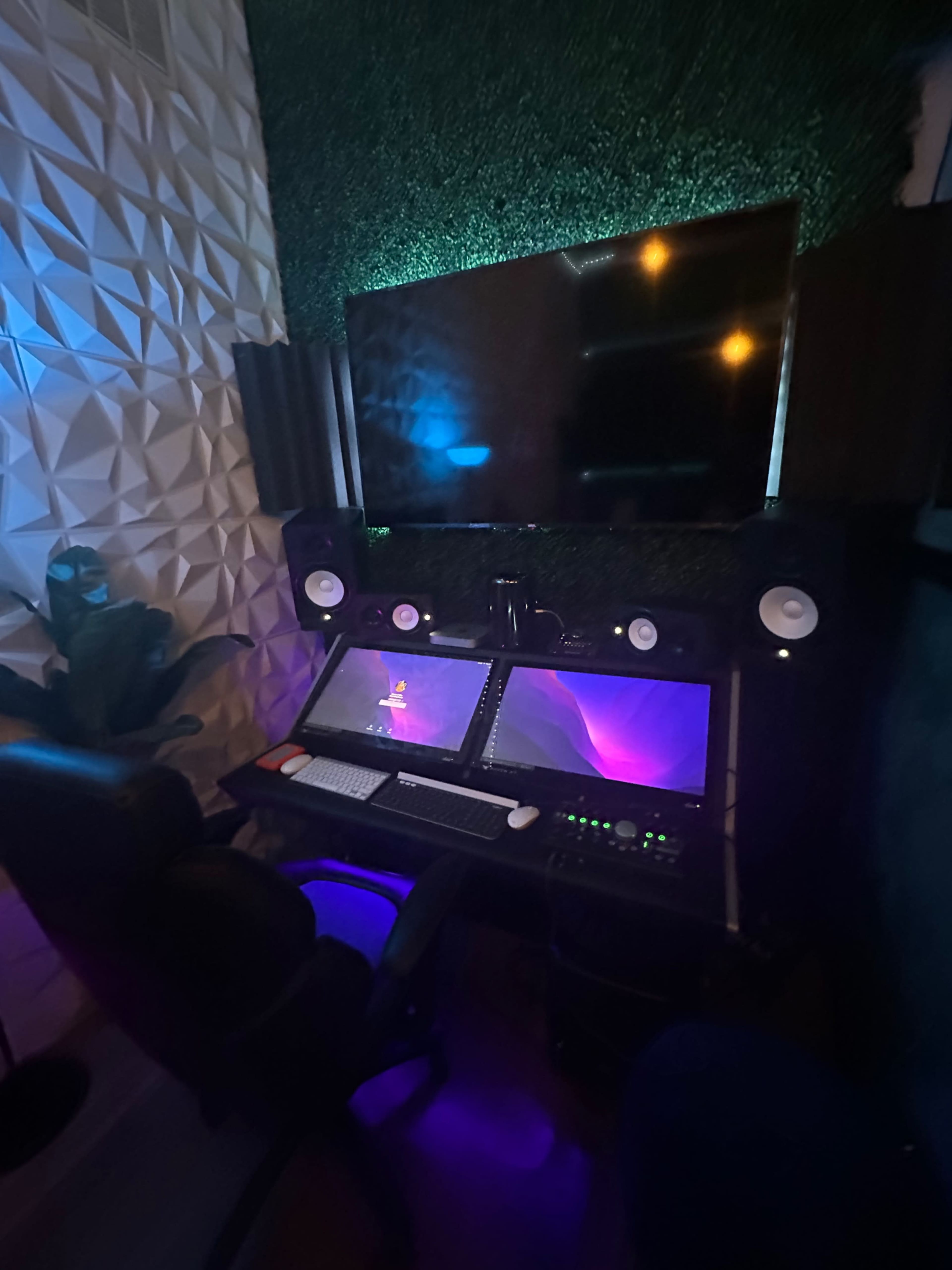 A recording studio setup featuring two computer monitors, studio speakers, and a large screen mounted on a textured wall.