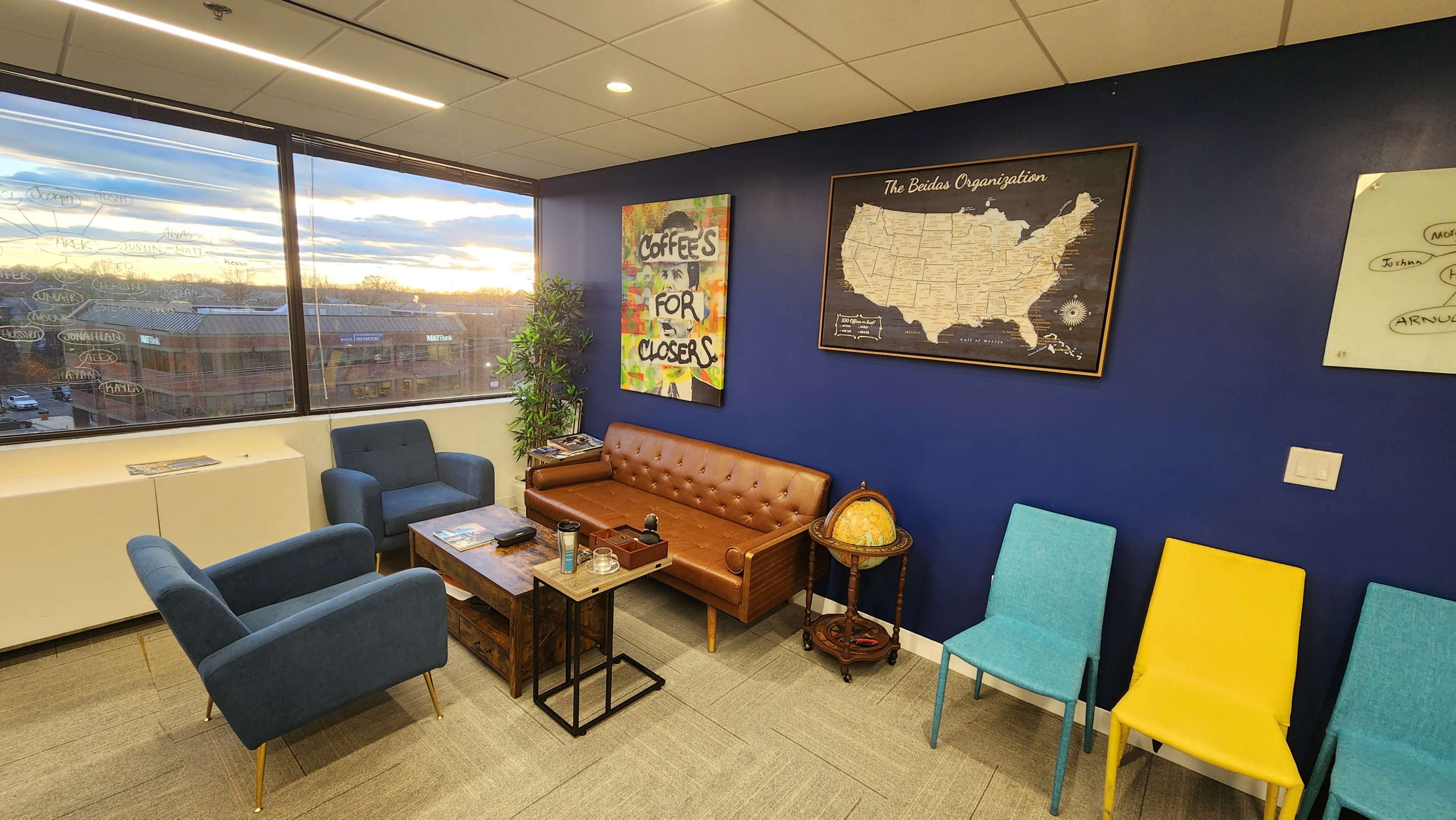 An office lounge area with a brown leather sofa, blue armchairs, a wooden coffee table, a globe, and colorful yellow chairs against a dark blue wall with a map and artwork.