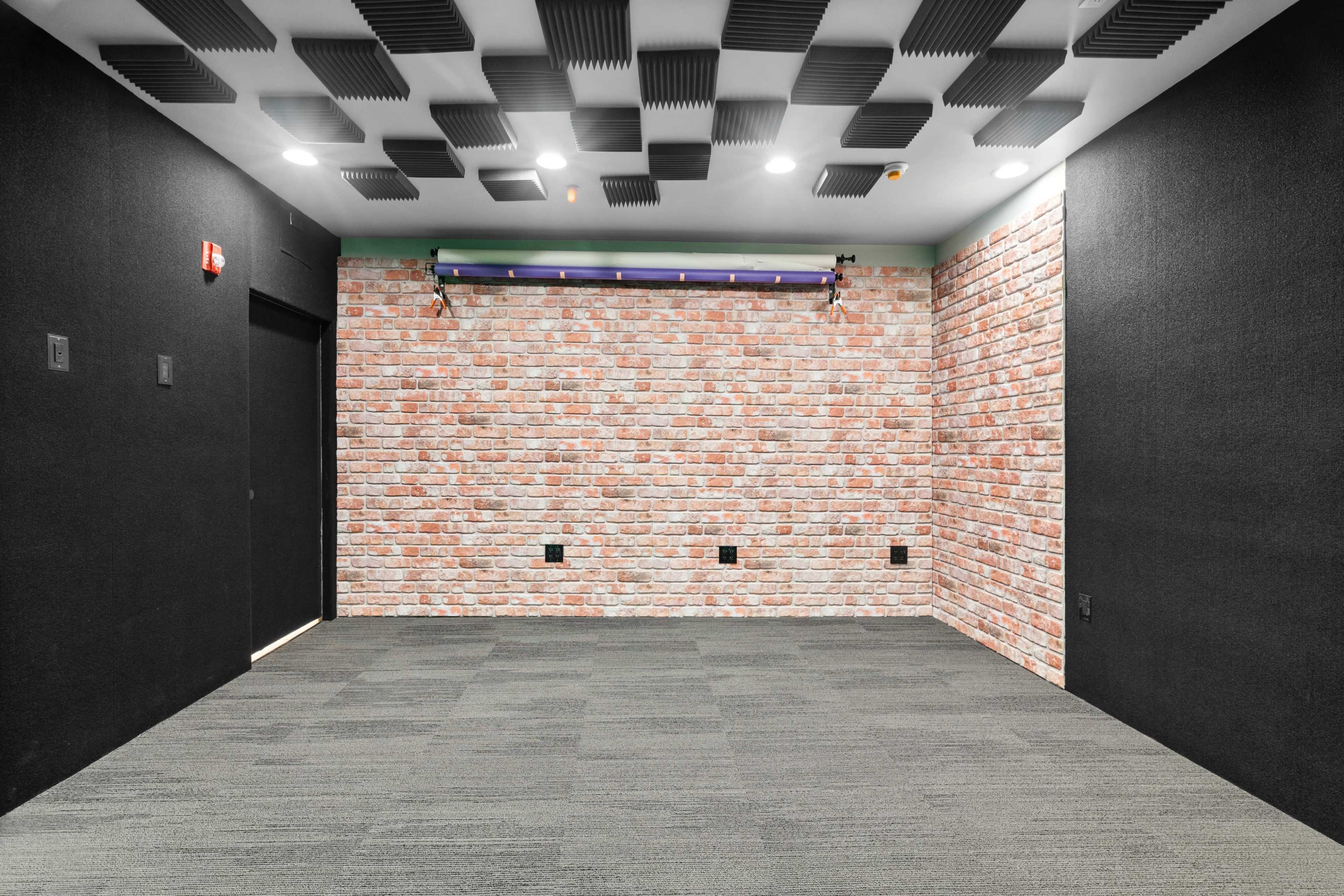 A small soundproof room with black walls, a brick-patterned accent wall, and a carpeted floor.