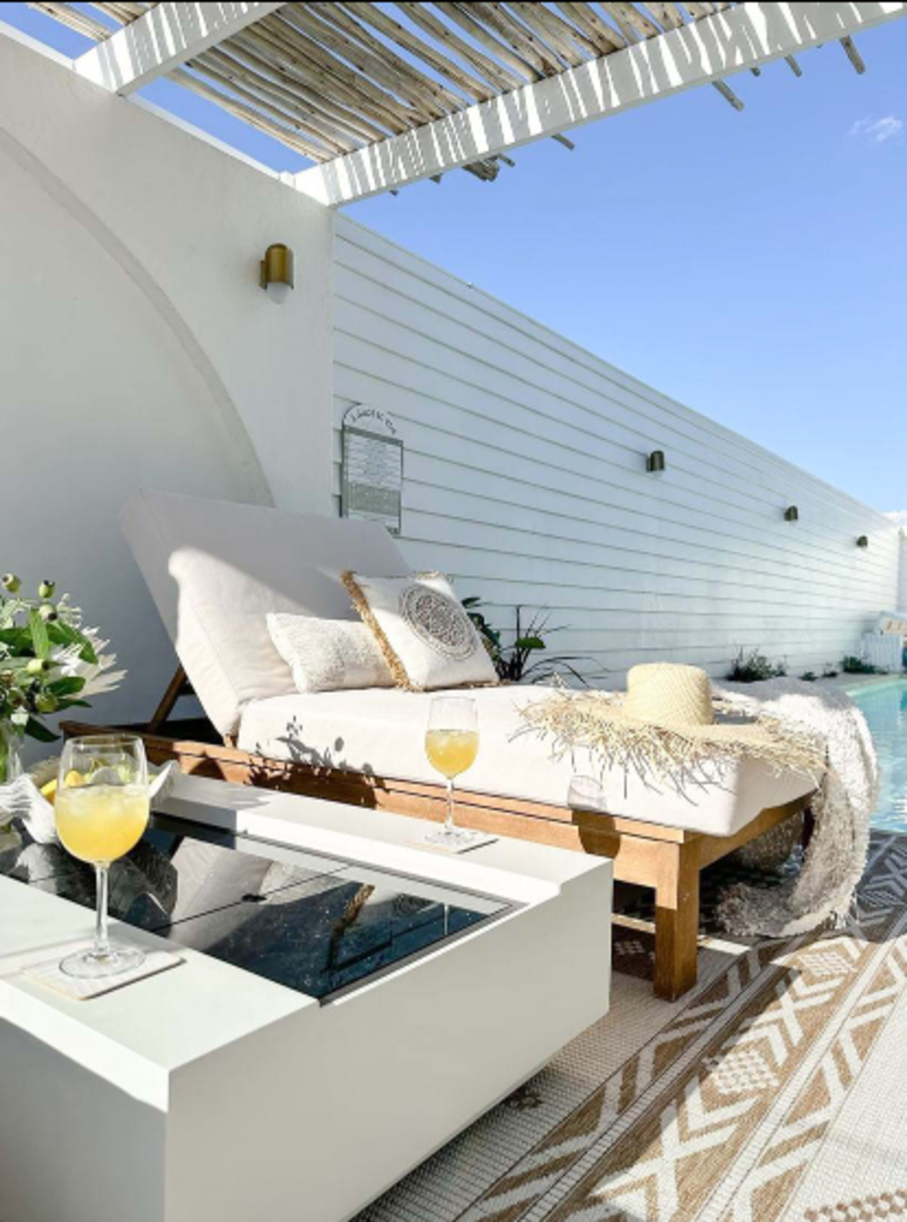 A chaise lounge with decorative pillows is positioned next to a pool, alongside a coffee table with two glasses of yellow beverages.