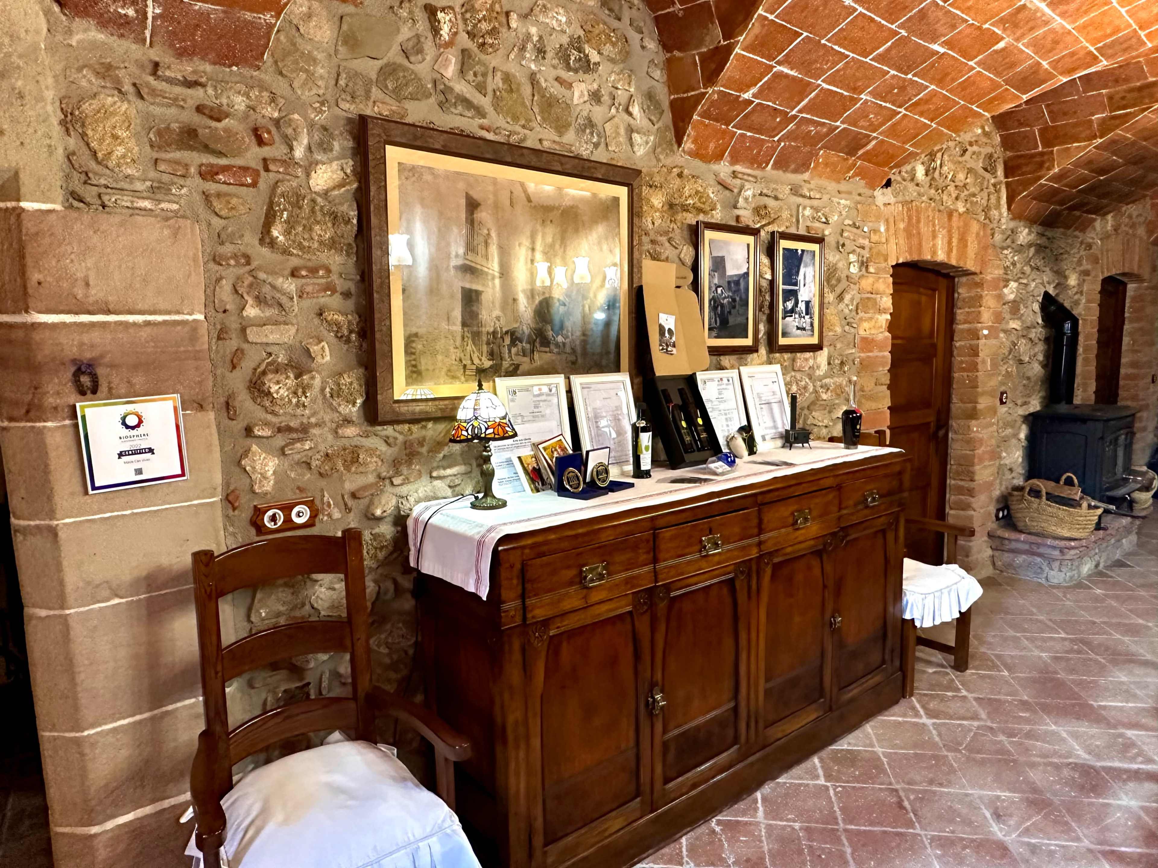 The image shows a rustic room with stone walls, featuring a wooden sideboard adorned with framed photographs and memorabilia, alongside a chair and a stove.