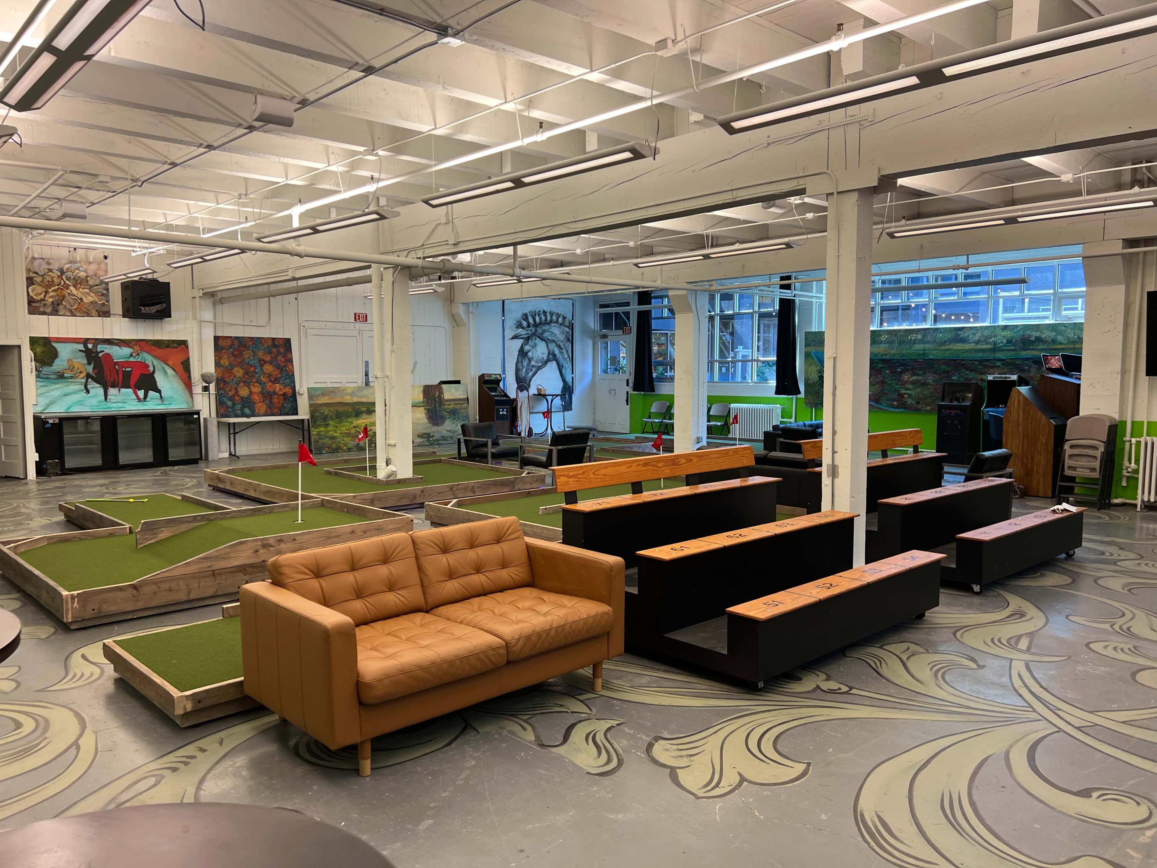 The image shows an indoor space featuring a mini-golf area with green turf, wooden seating, and large windows providing natural light.