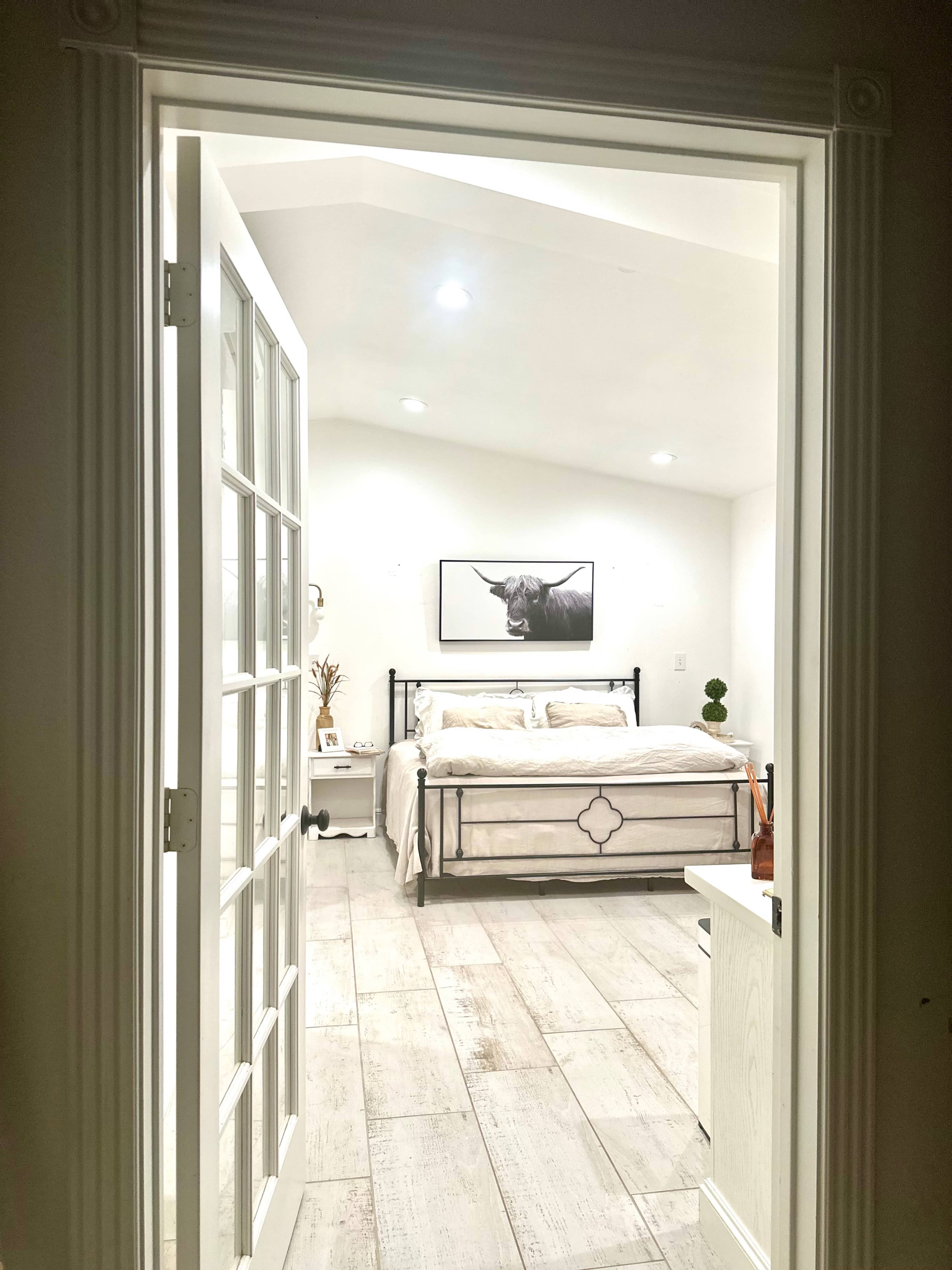 A view of a bedroom through an open door, featuring a bed with white bedding and a framed picture of a bull on the wall.