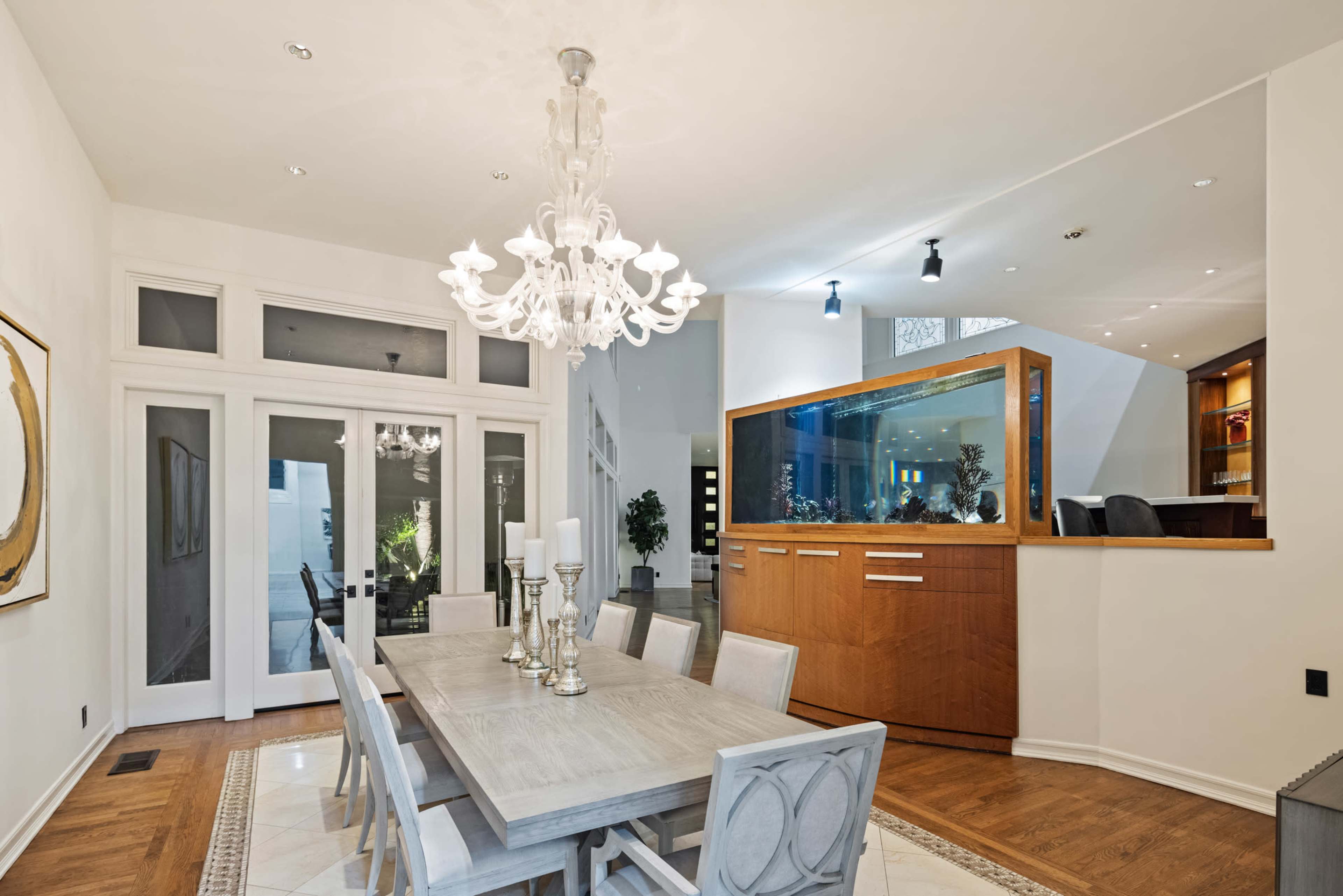 The image shows a well-lit dining area featuring a large wooden table with chairs, a chandelier above, and an aquarium in the background.