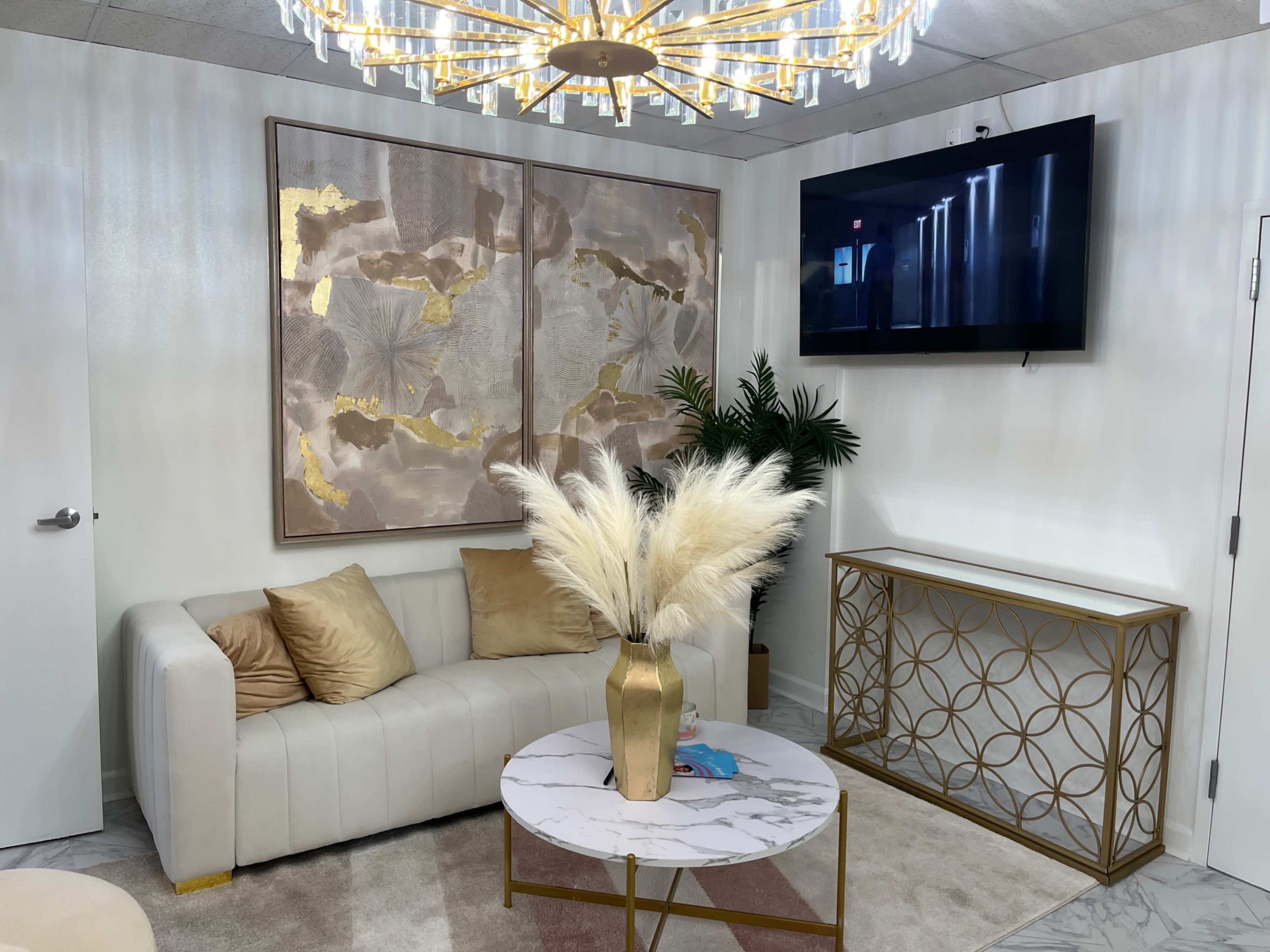A modern waiting area features a white leather sofa, a sleek gold-accented coffee table with a marble top, and a decorative arrangement of dried plants, alongside a large wall art piece and a flat-screen TV.