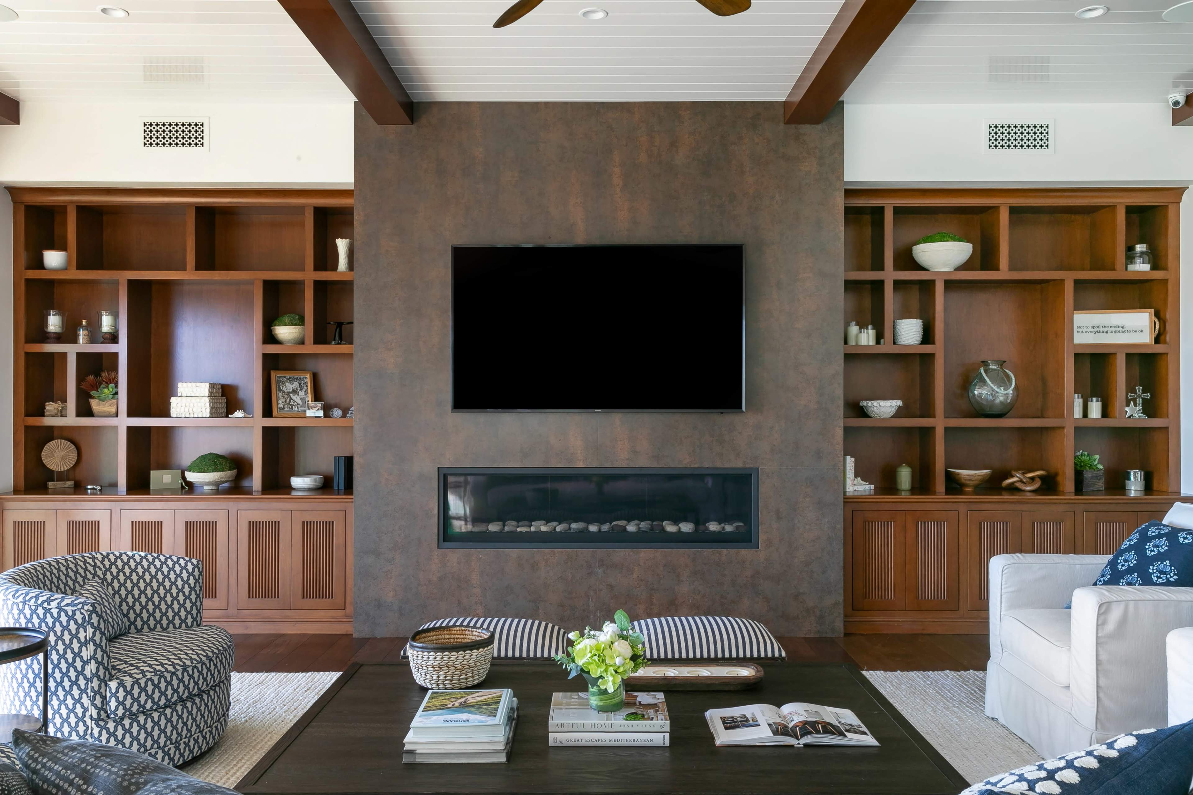 A modern living room features a central fireplace below a large flat-screen television, flanked by built-in wooden shelves filled with decorative items and books.