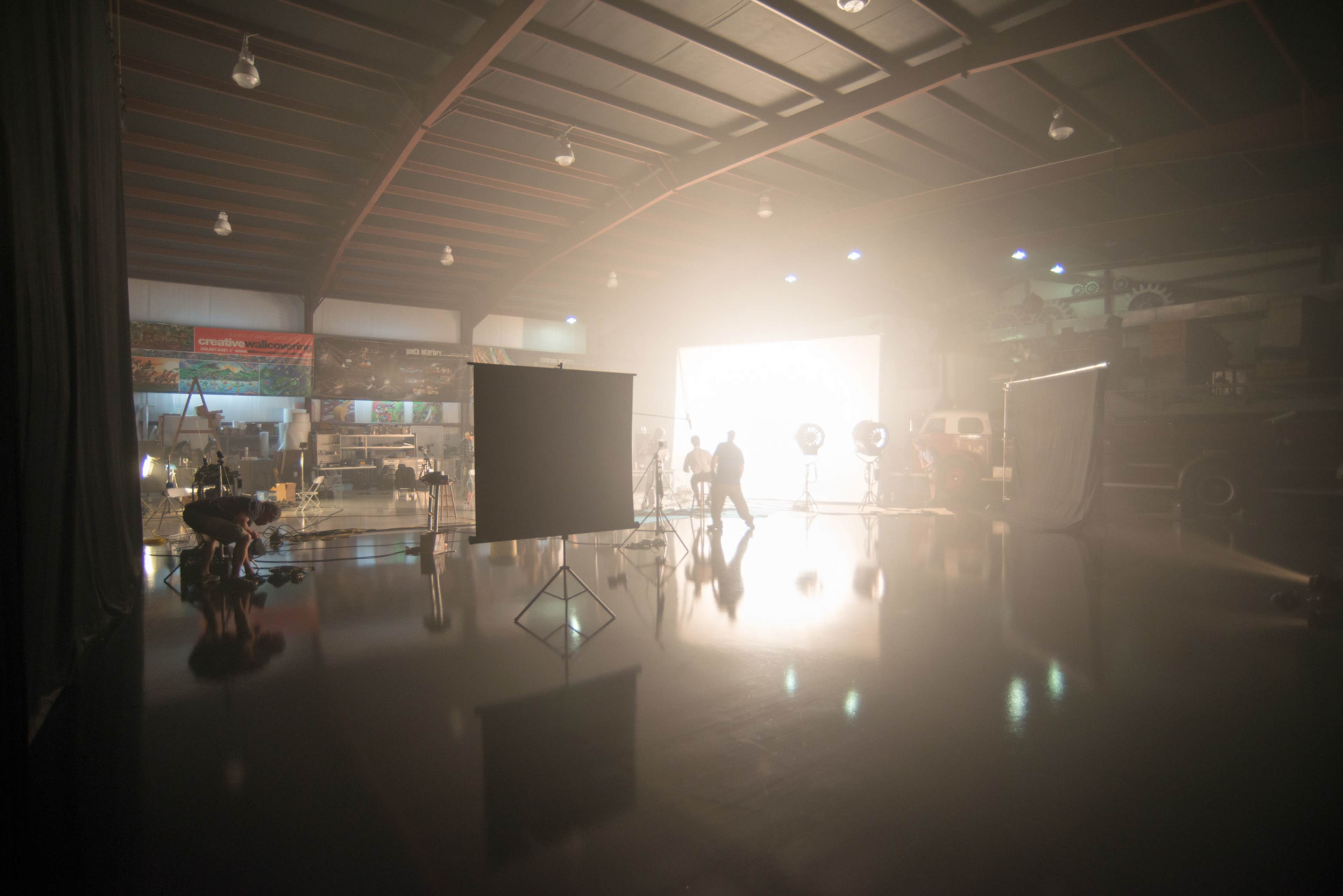 A large film studio is set up with bright lights, reflective surfaces, and various filming equipment.