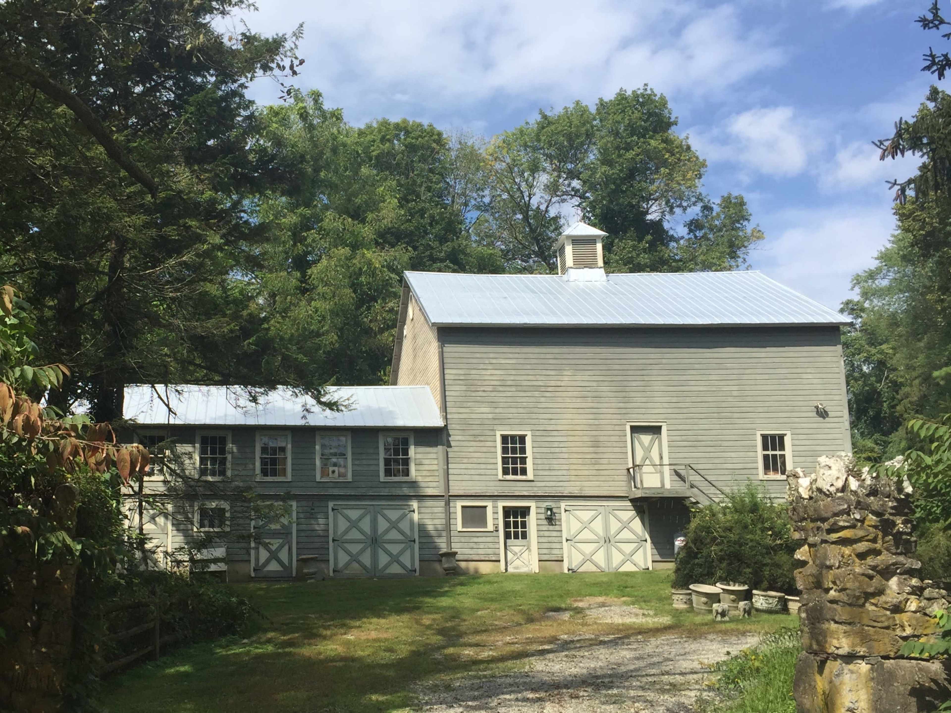 A two-story gray wooden barn with a metal roof and several windows, surrounded by trees and a gravel driveway.