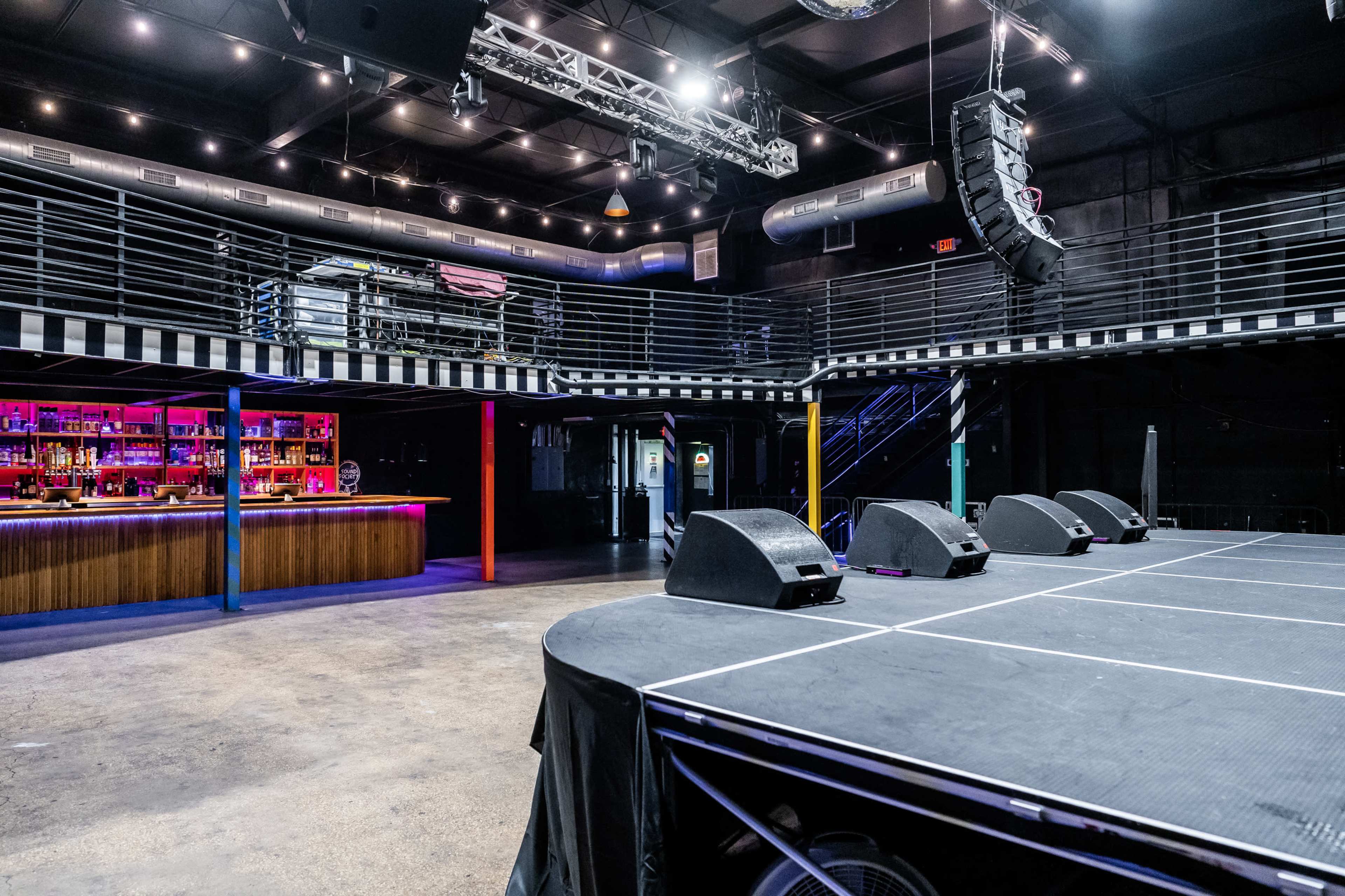 An empty music venue with a stage in the foreground and a bar area illuminated in the background, featuring a balcony above.