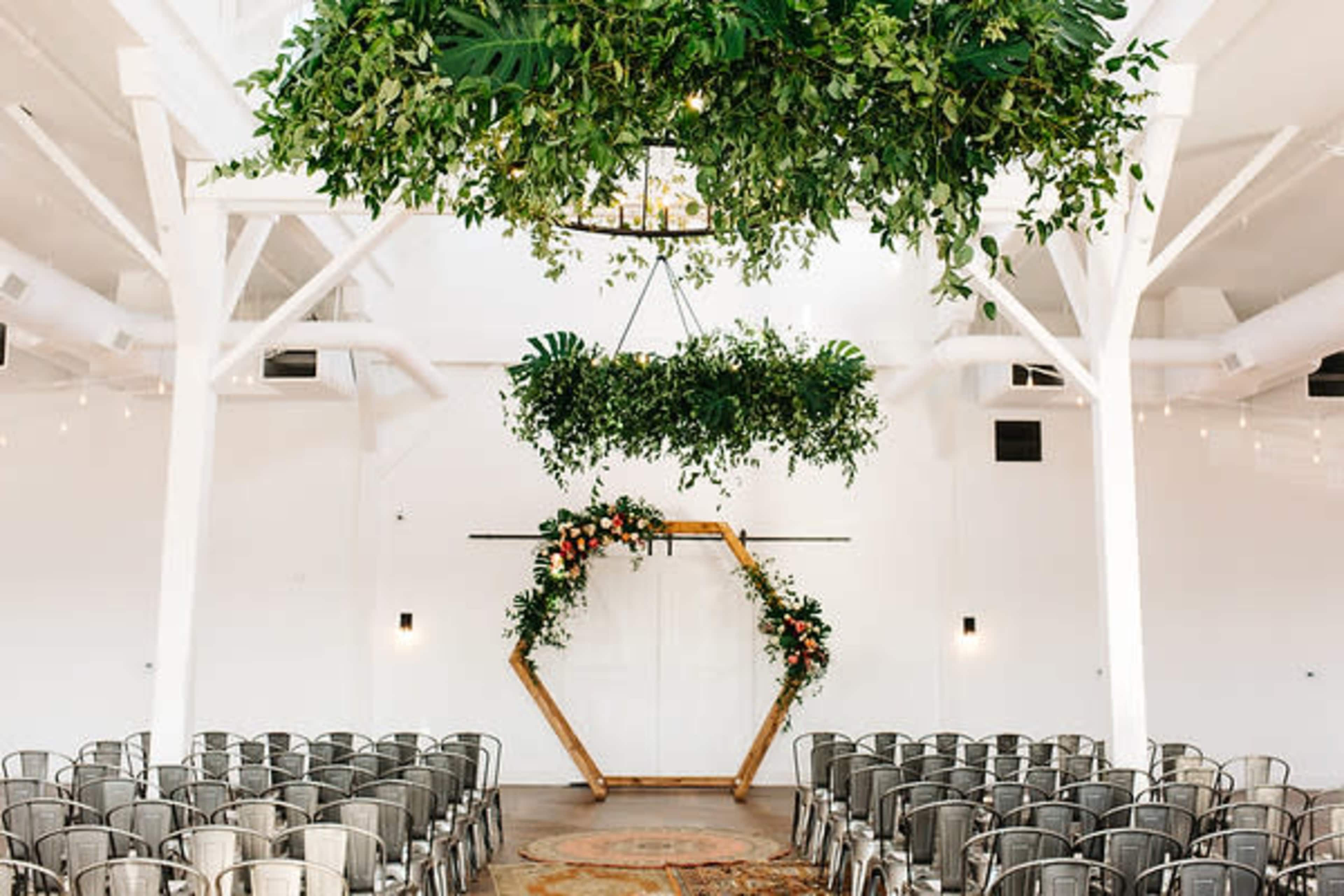 A wedding ceremony space with a geometric wooden arch adorned with greenery and flowers, surrounded by rows of metal chairs under hanging plants and lights.