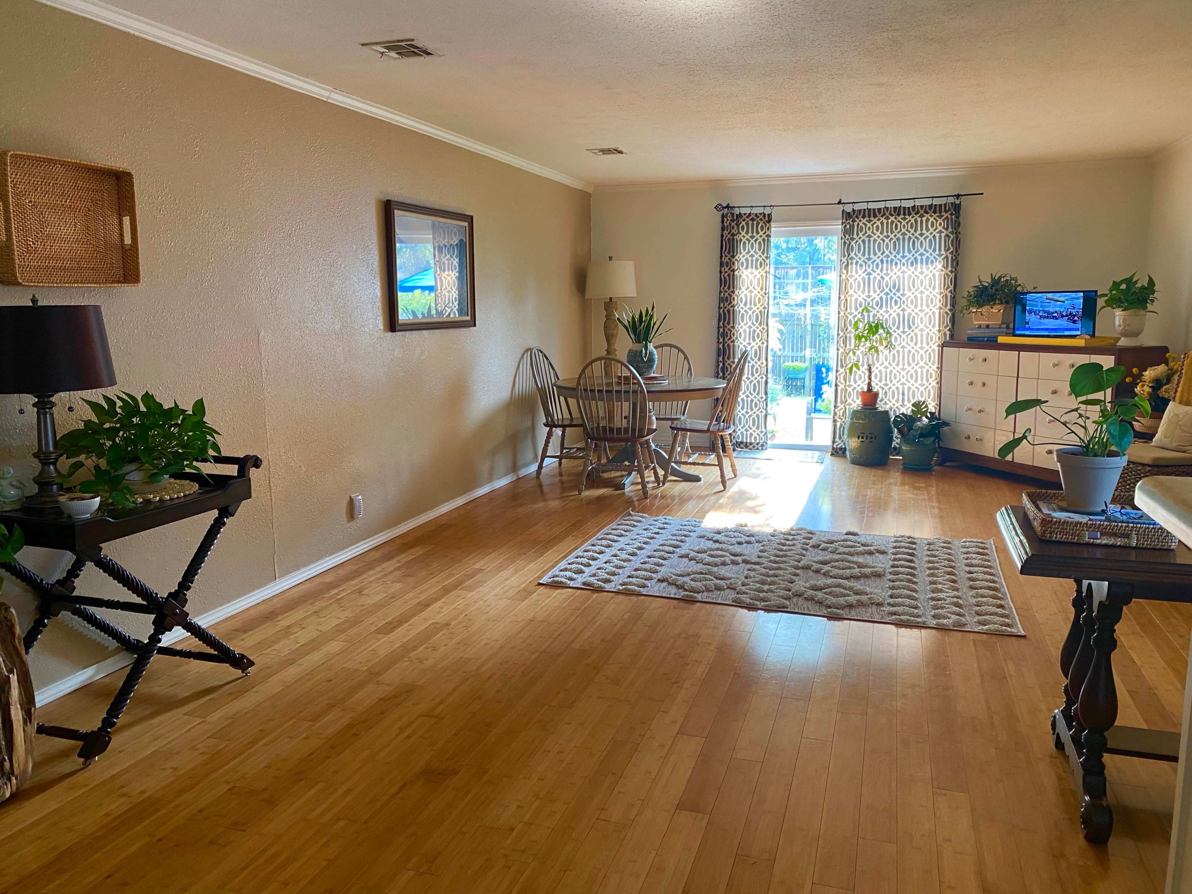 The room features bamboo flooring, a dining area with a table and chairs, a small seating area with a lamp and plants, and large windows leading to a patio.