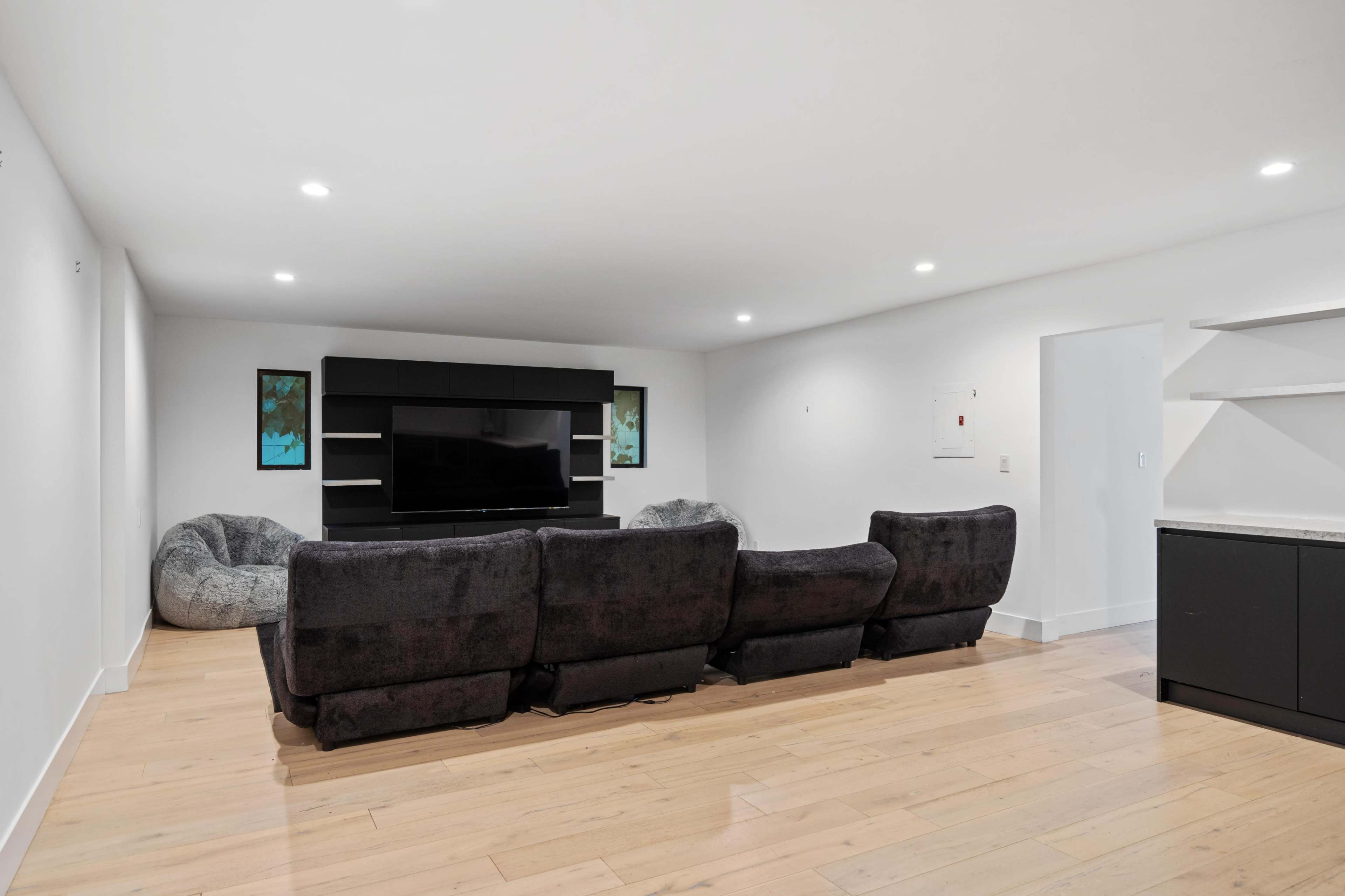 A modern living room with a black sectional sofa facing a large flat-screen TV on a wall, alongside two beanbag chairs.