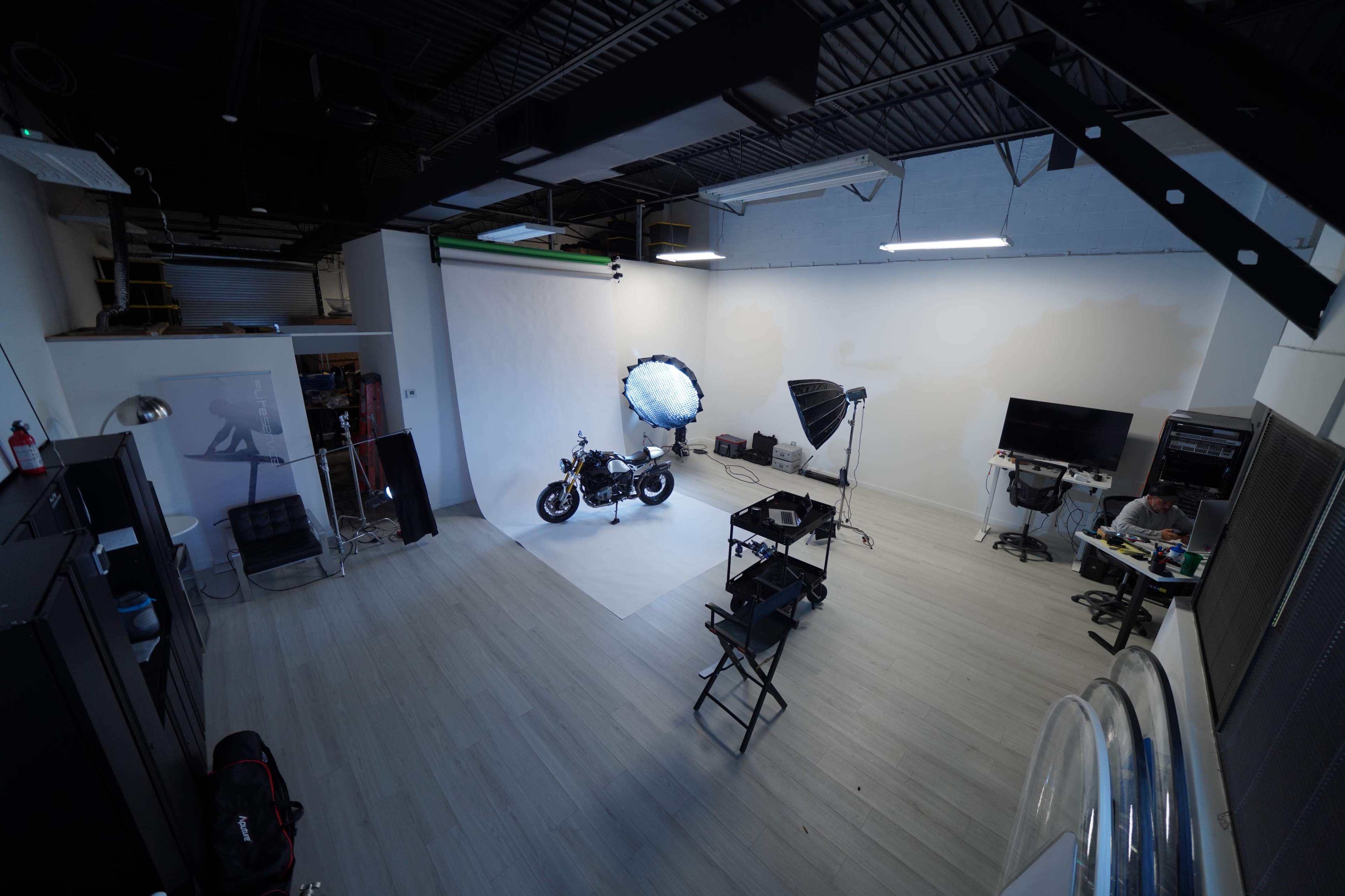 The image shows a well-lit photography studio featuring a motorcycle on a white backdrop, along with several lighting equipment and a work area.