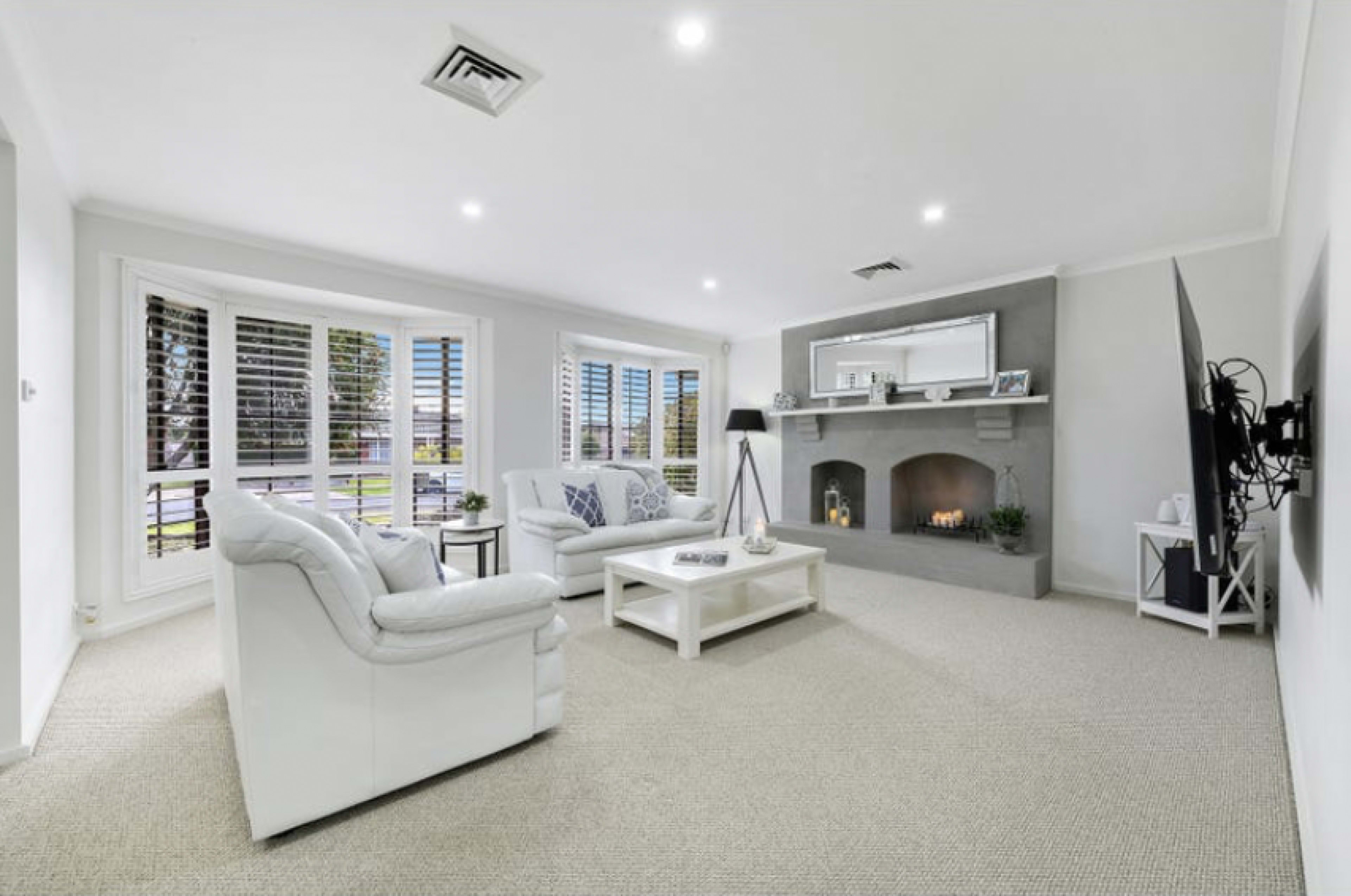 The image shows a modern living room with white furniture, a gray fireplace, and large windows featuring plantation shutters.