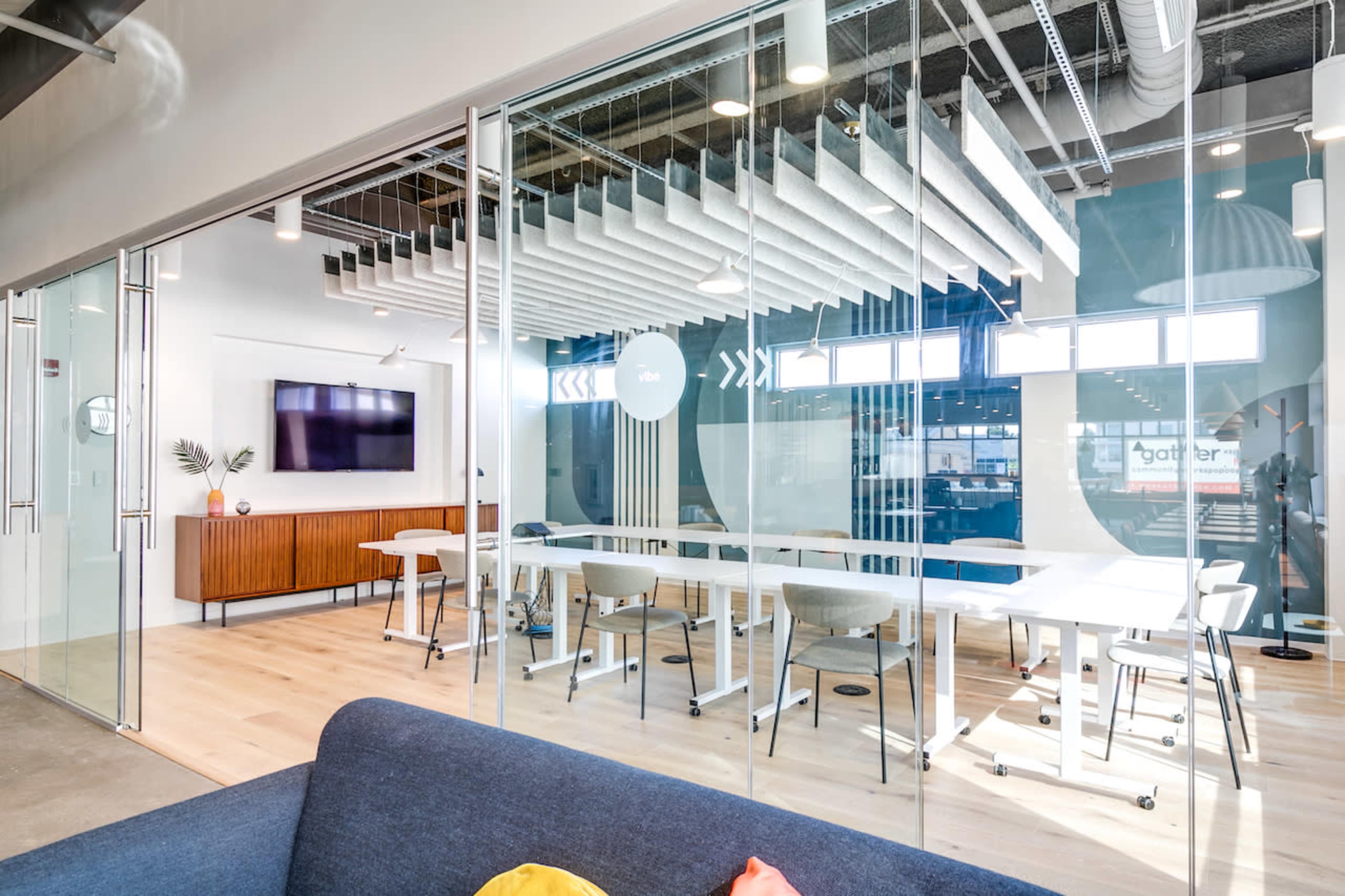 A modern office conference room features a large glass wall, white tables, and a wall-mounted television.