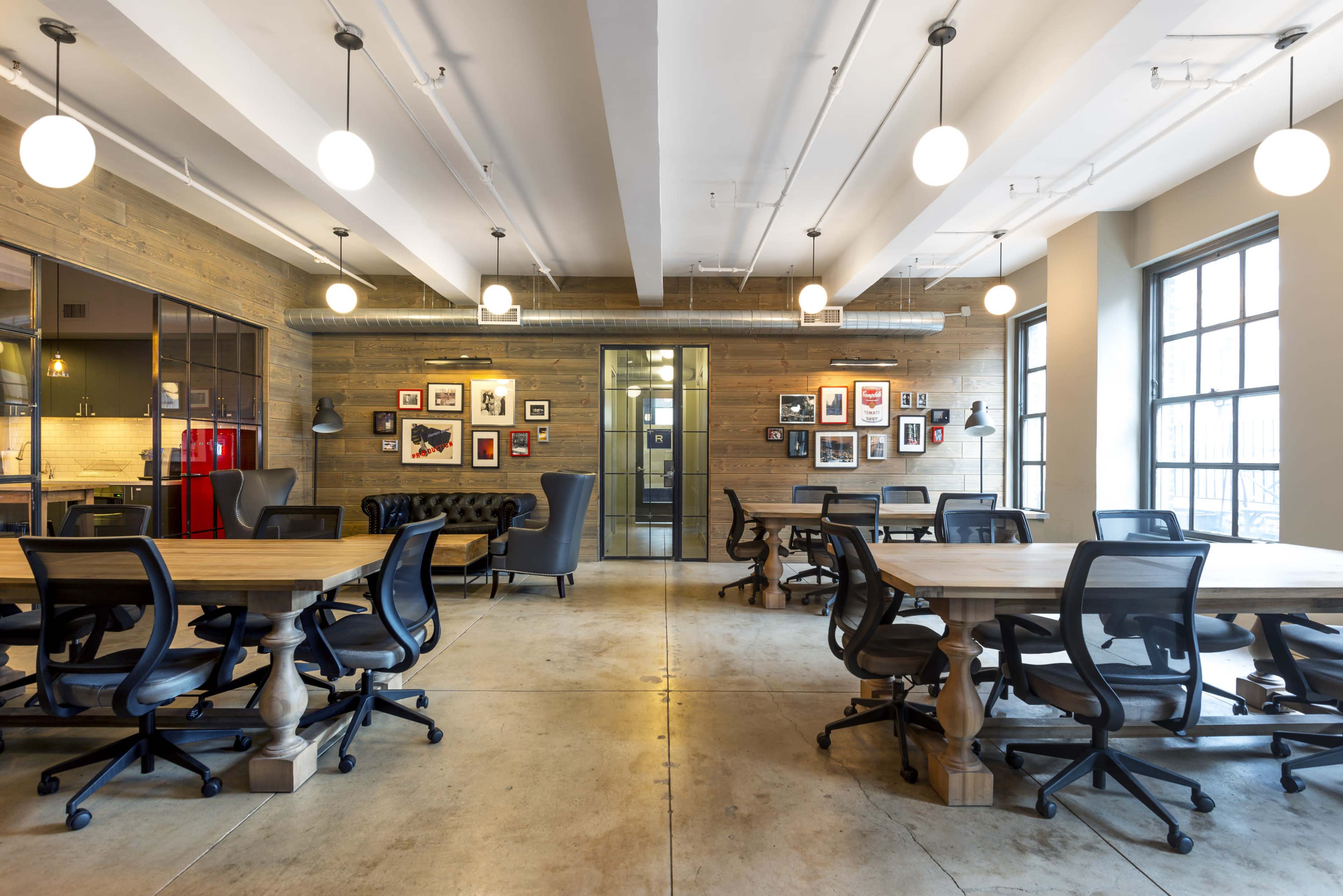 The image shows a modern office space with wooden walls, several tables, and black chairs, illuminated by round pendant lights.