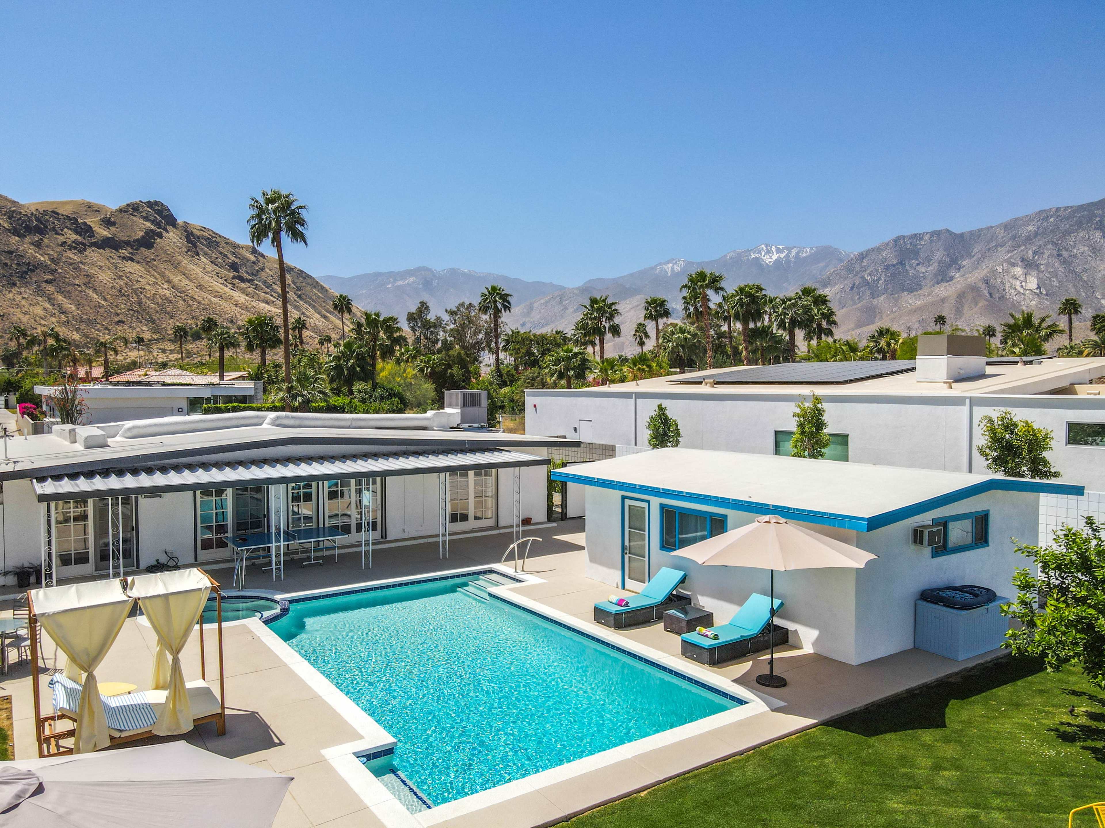 A modern, white house with a swimming pool surrounded by palm trees and mountains in the background.