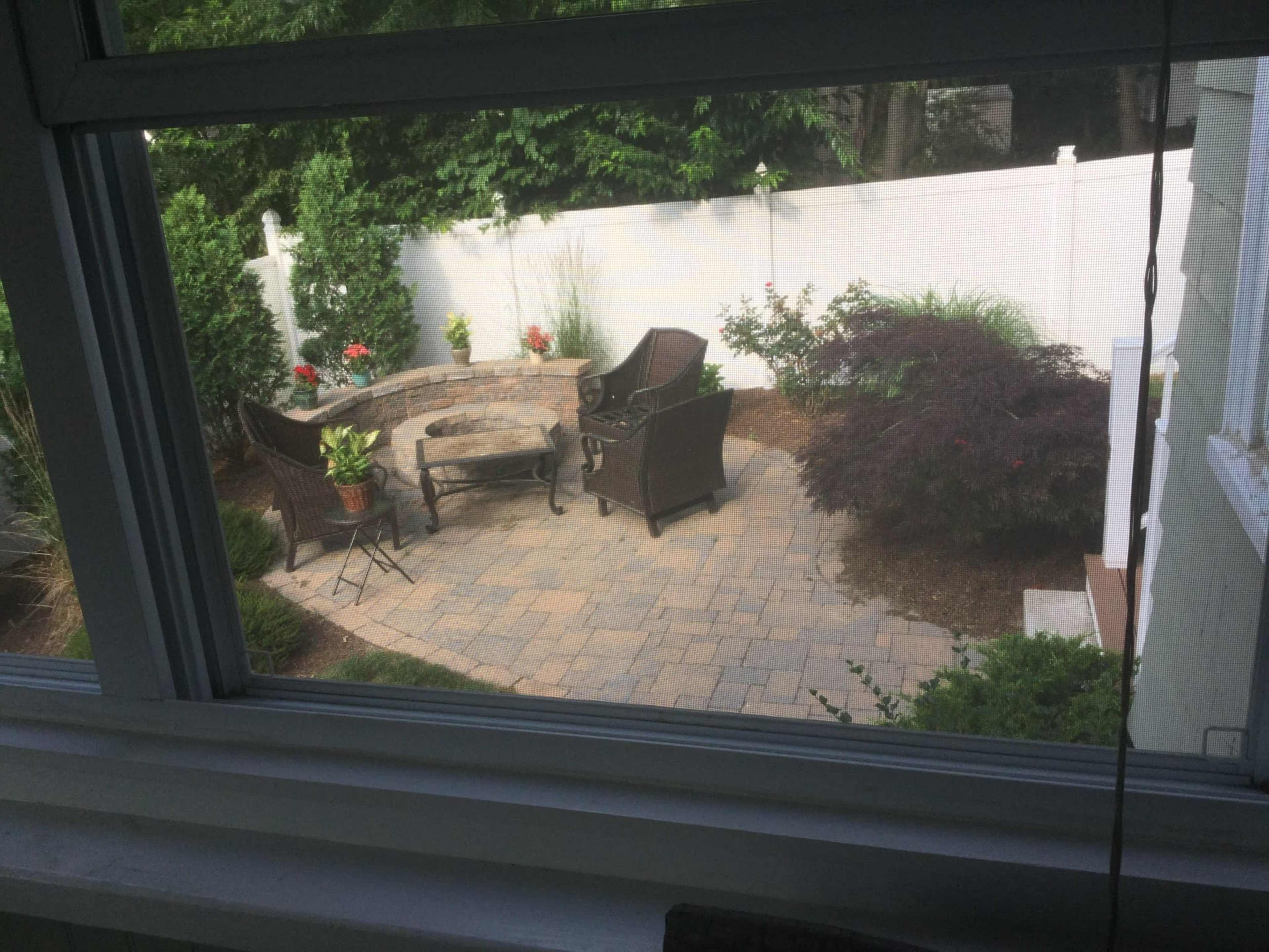 A stone patio features two wicker chairs and a small table surrounded by landscaped greenery and a white fence.