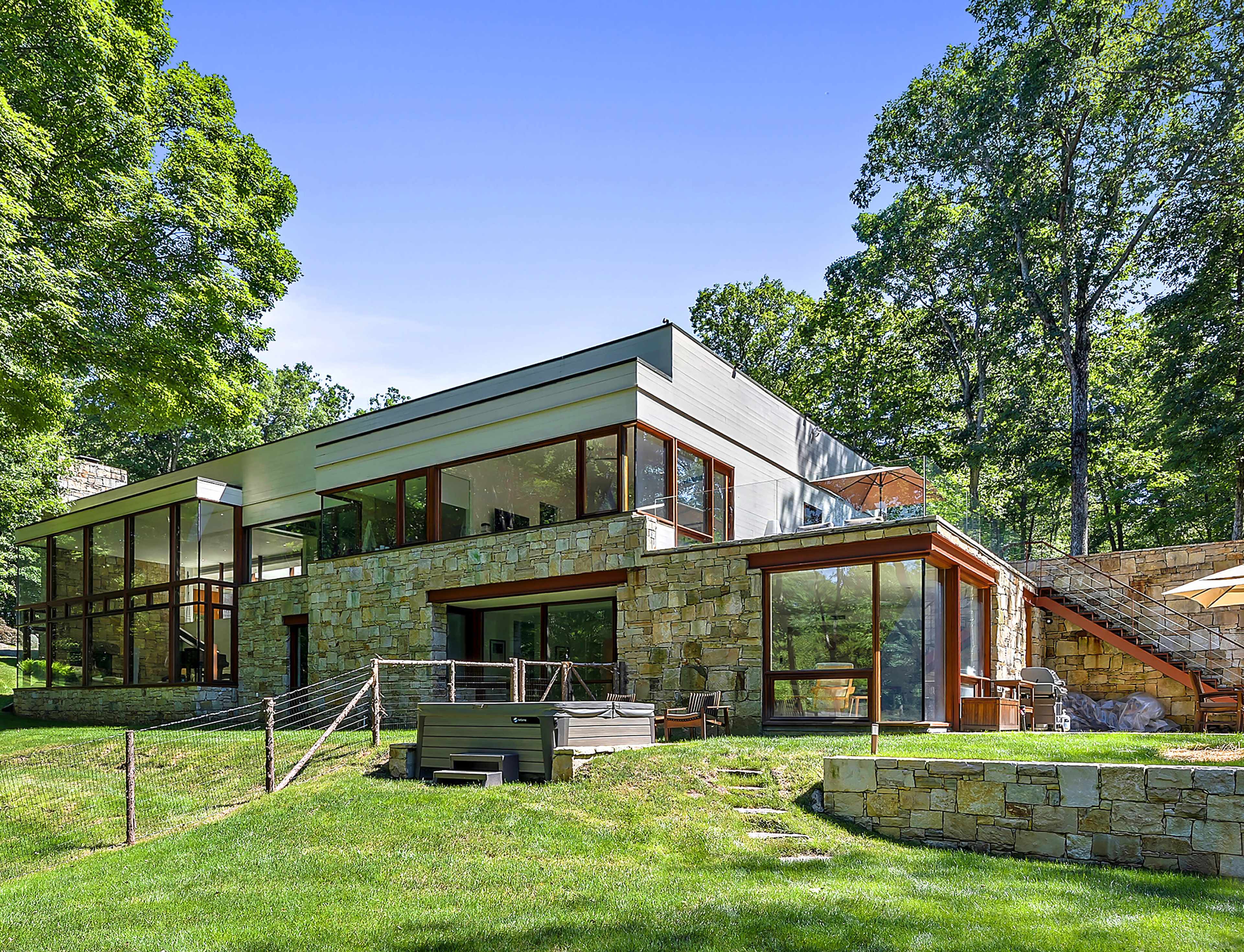 The image shows a modern two-story stone and glass house set in a wooded area, featuring landscaped grounds and a patio.