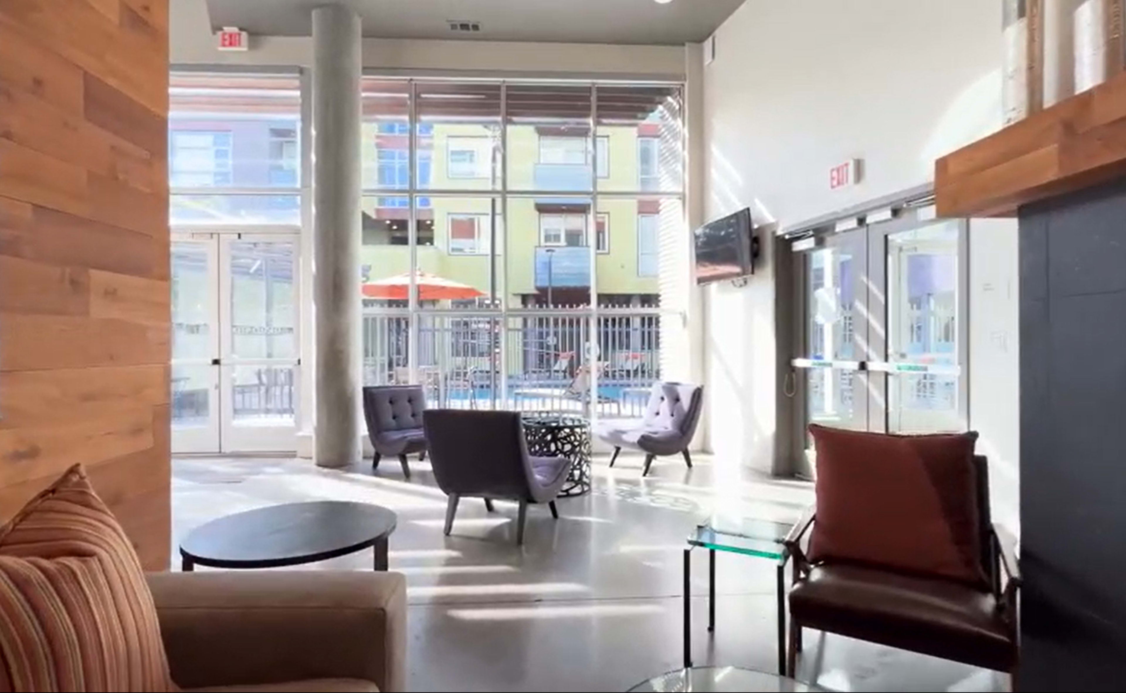 The image shows a modern lounge area with seating, large windows, and a view of an outdoor space with patio umbrellas.