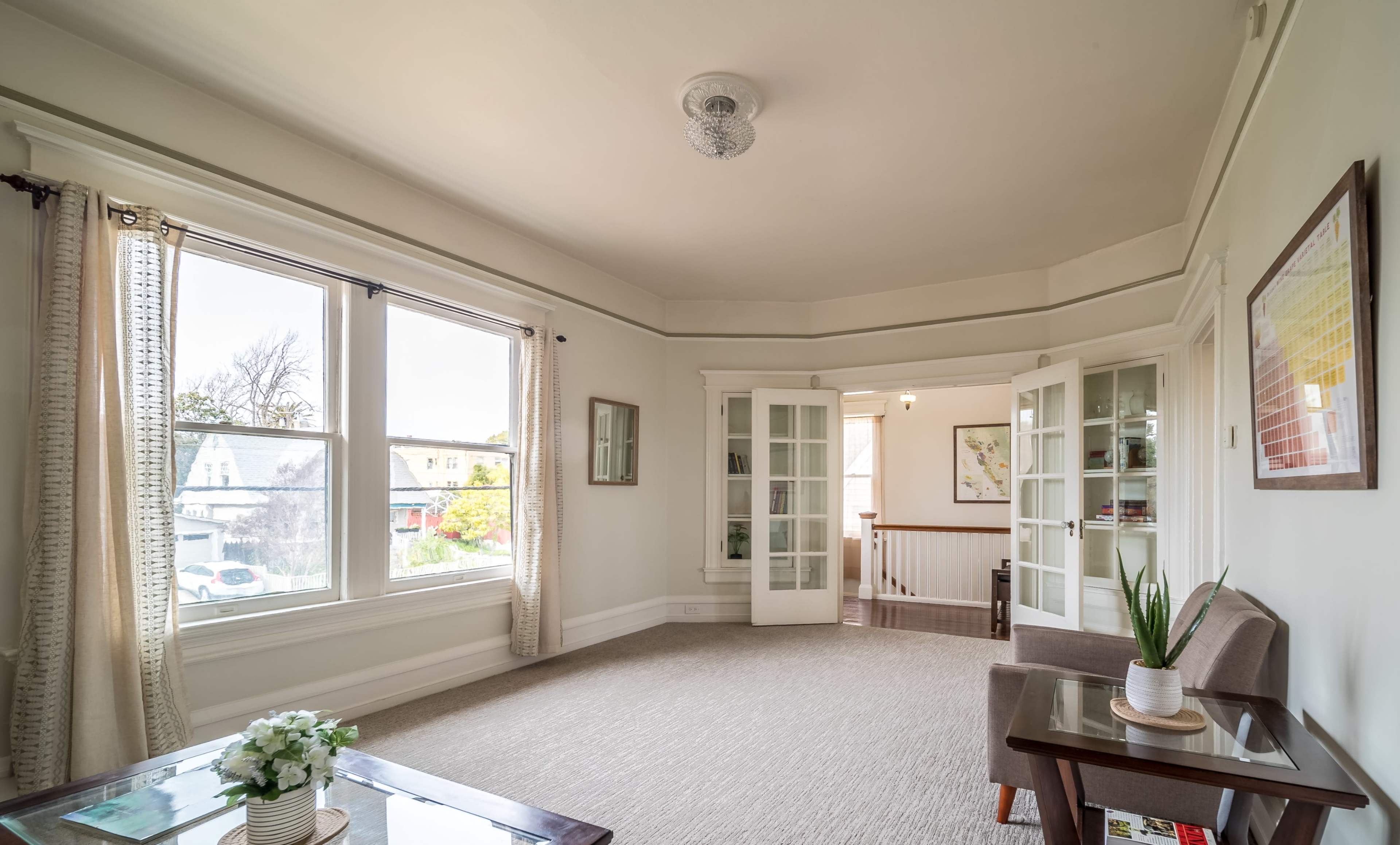 A well-lit room with large windows, carpeted flooring, and a small table is furnished with a chair and decorative plants.