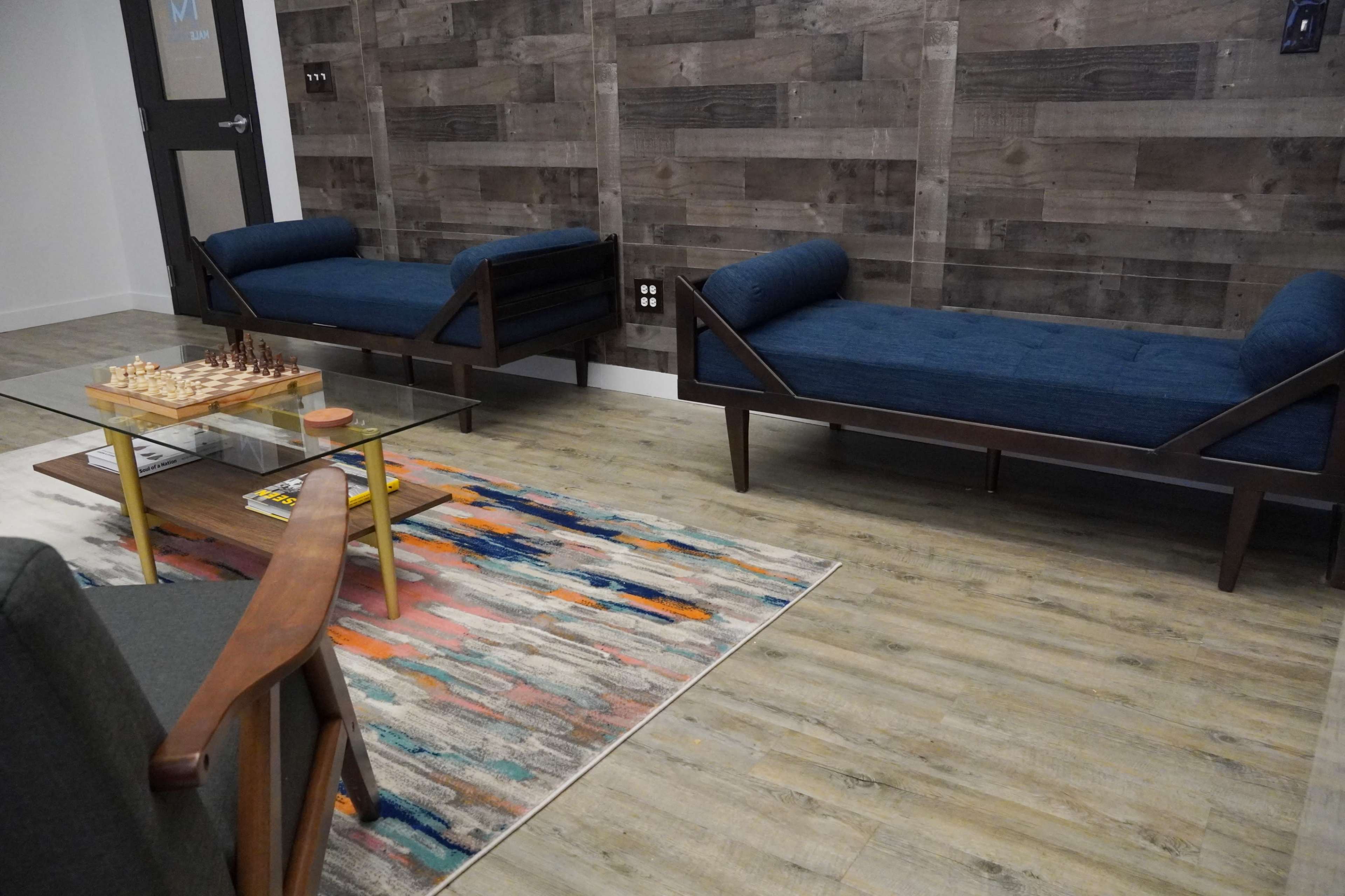 A modern lounge area with two blue upholstered couches, a glass coffee table on a colorful rug, and a chessboard set on the table.