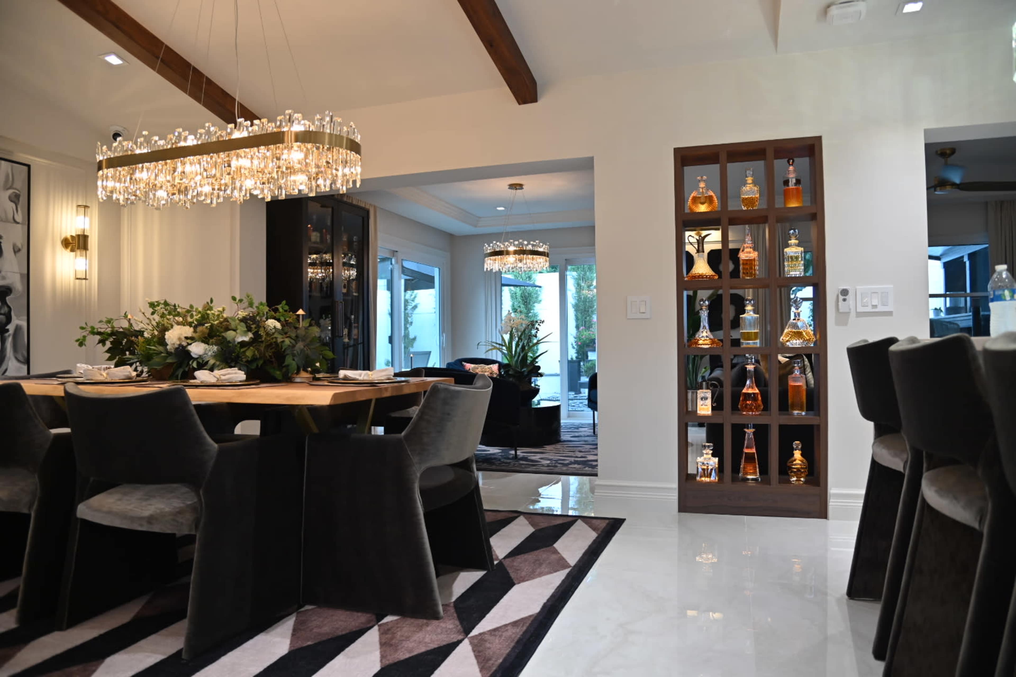 The image shows a modern dining area featuring a wooden table surrounded by dark chairs, a crystal chandelier overhead, and a display unit with illuminated bottles.