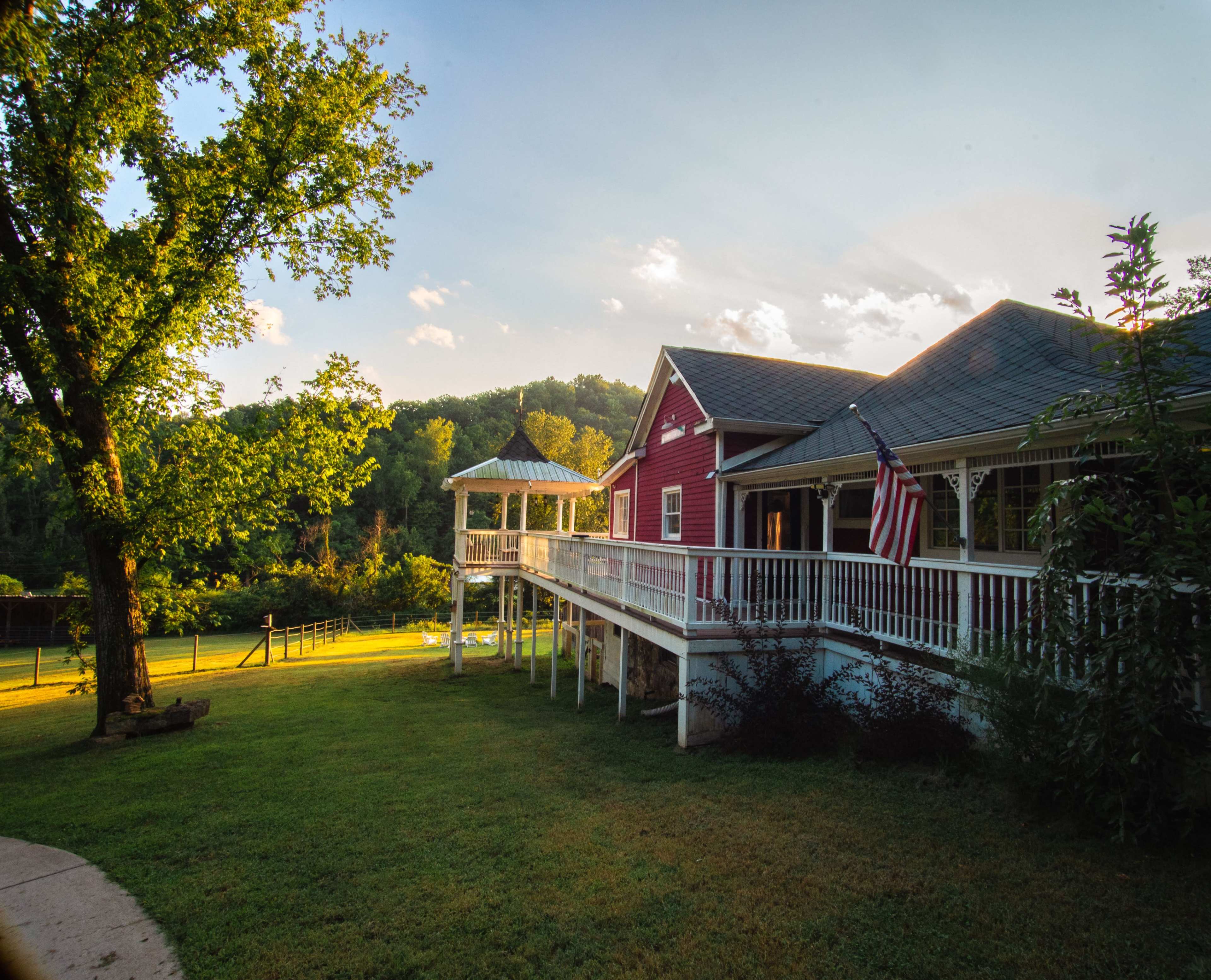 A red house with a porch and an American flag is situated in a green landscape with trees and hills in the background.